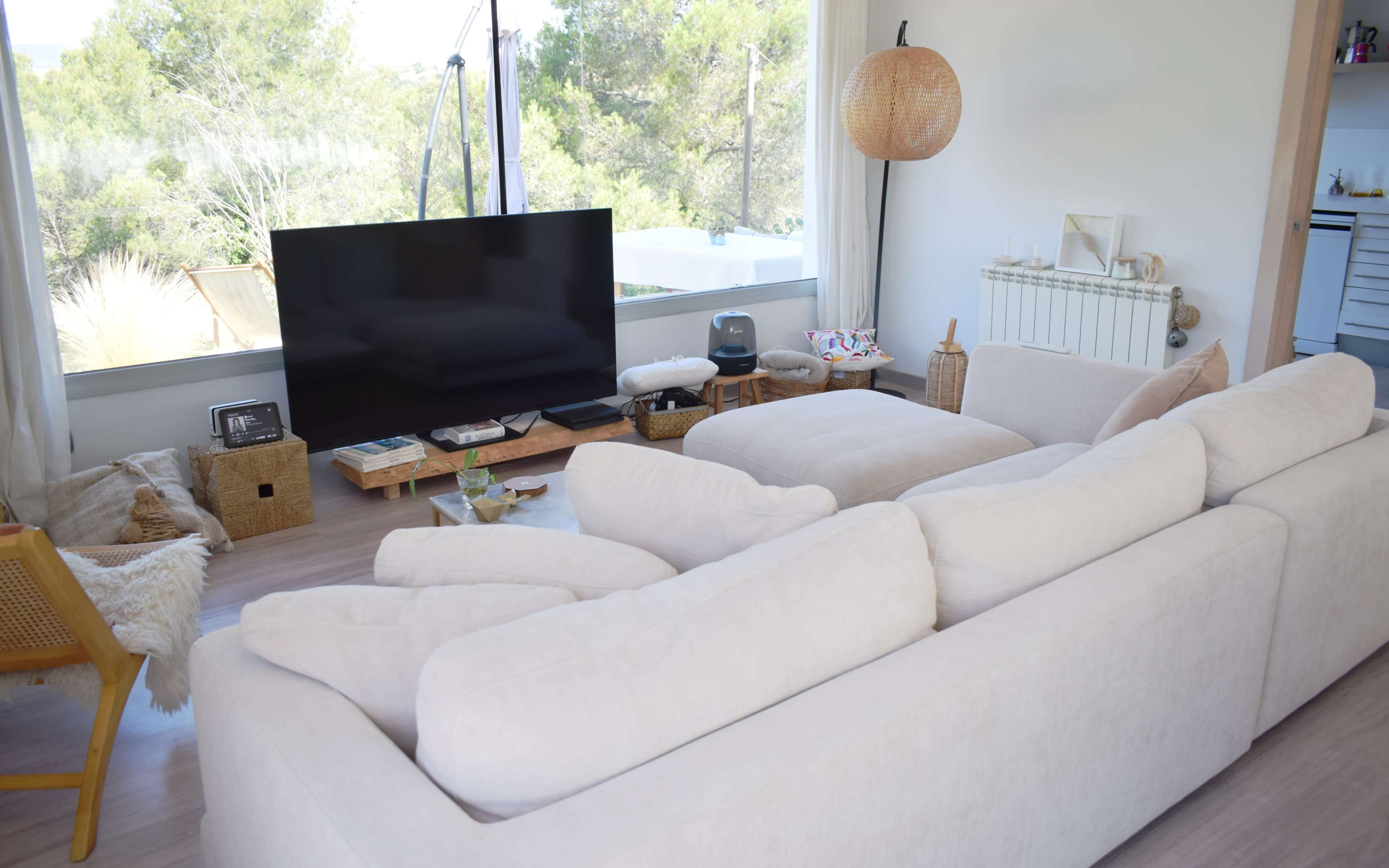 A light-colored sectional sofa faces a large television in a modern living room with expansive windows overlooking greenery.