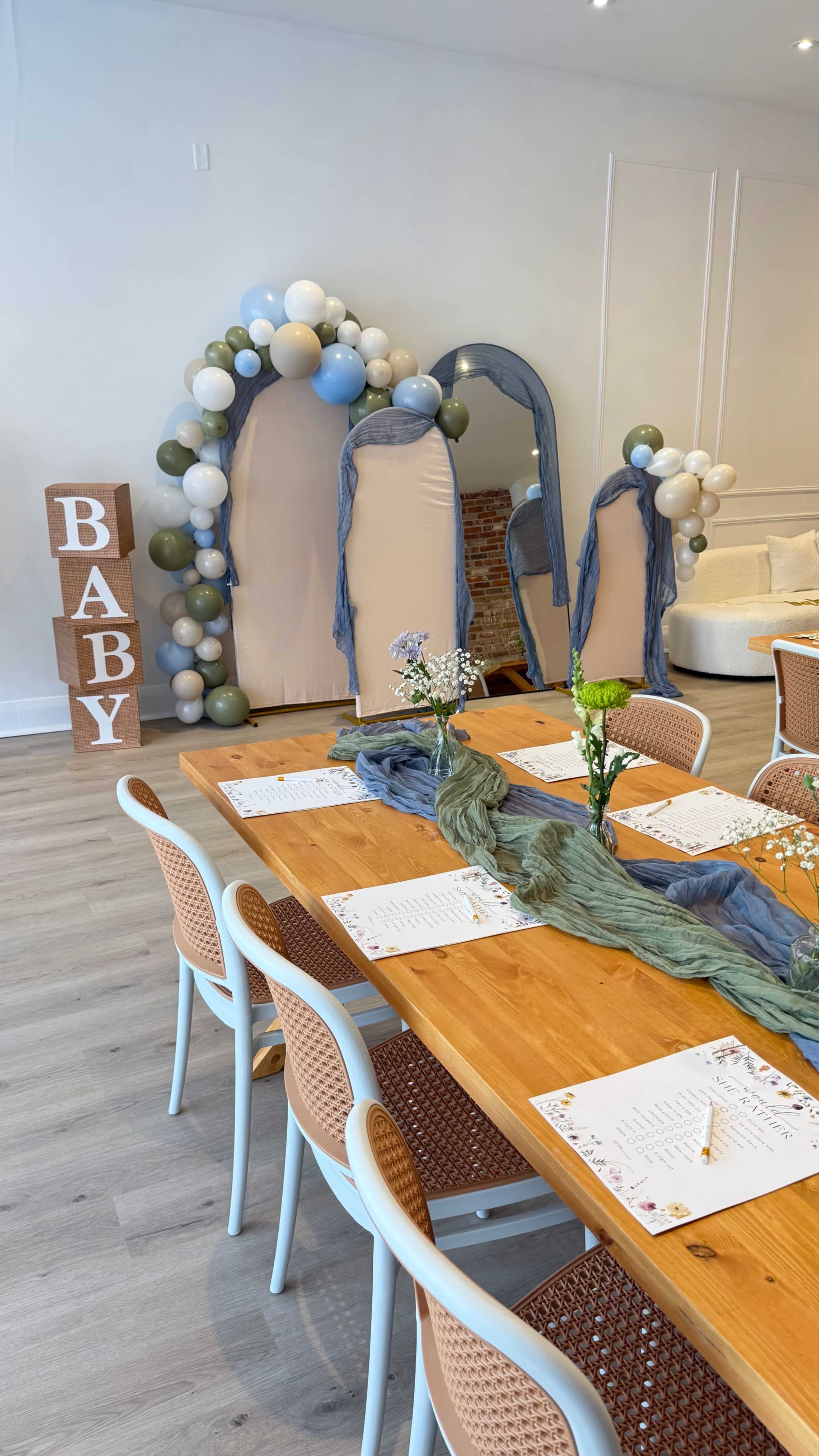 The image shows a decorated event space with a long wooden table set for a gathering, surrounded by balloon arches and "BABY" letters made of wooden blocks.