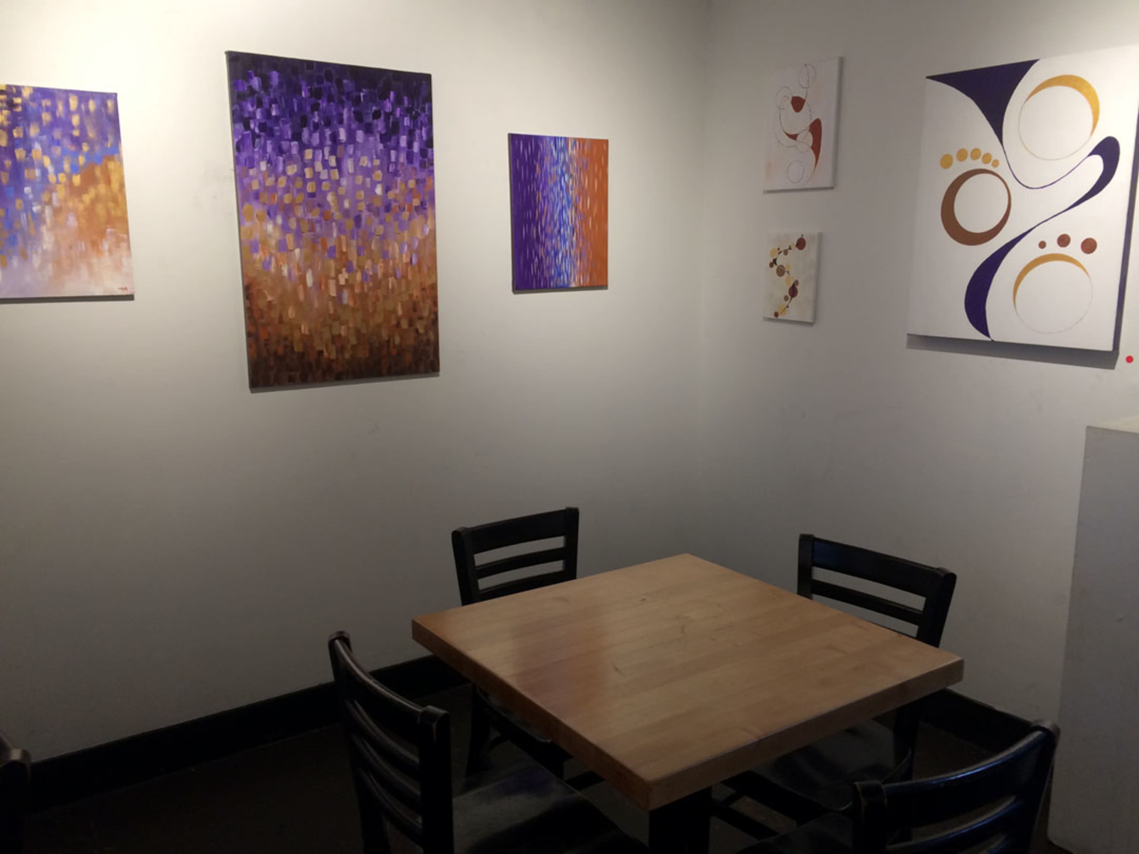 A small dining area with a wooden table surrounded by black chairs, featuring colorful abstract paintings on the walls.