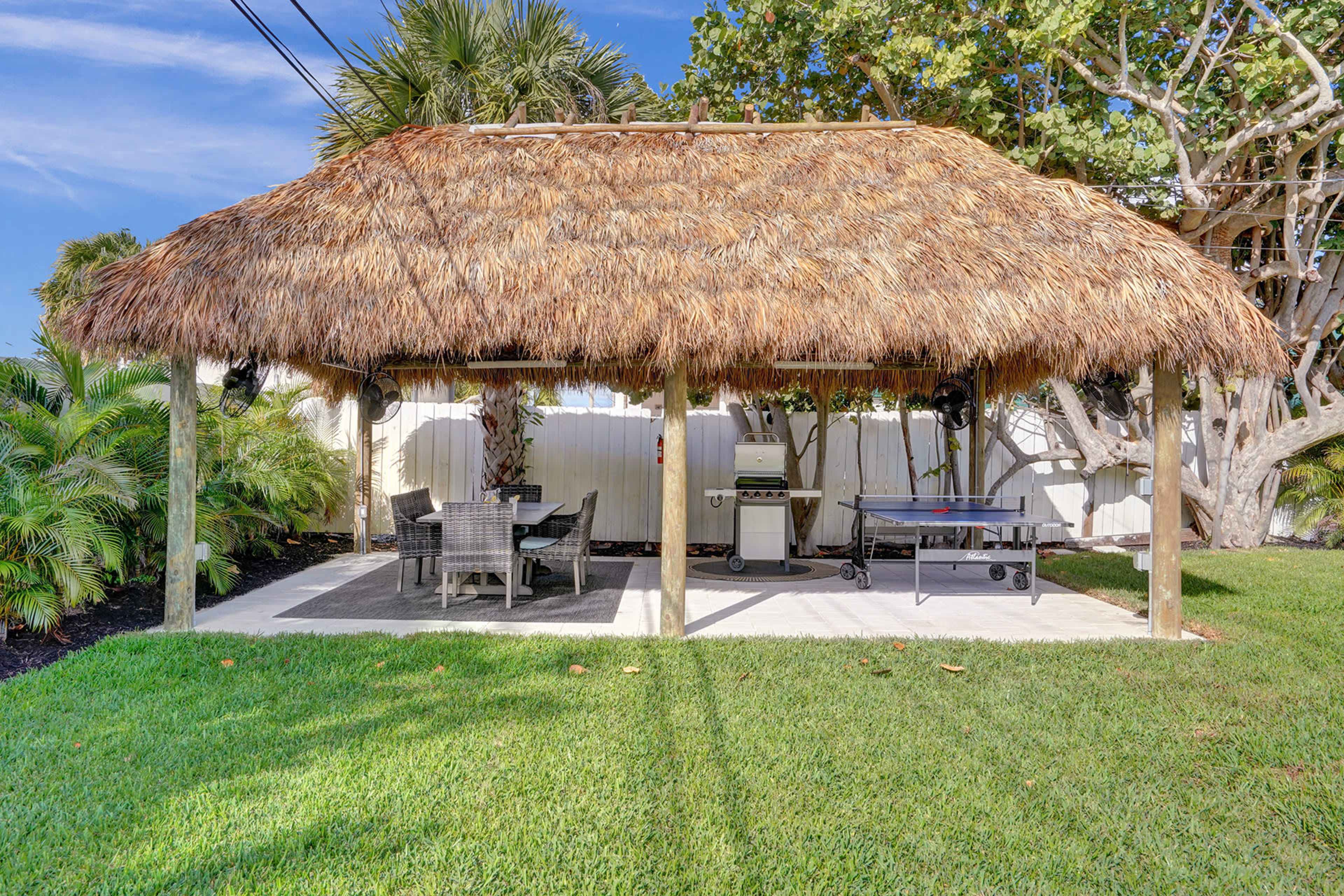 A thatched roof gazebo with a dining area and a grill is situated on a grassy lawn surrounded by palm trees.