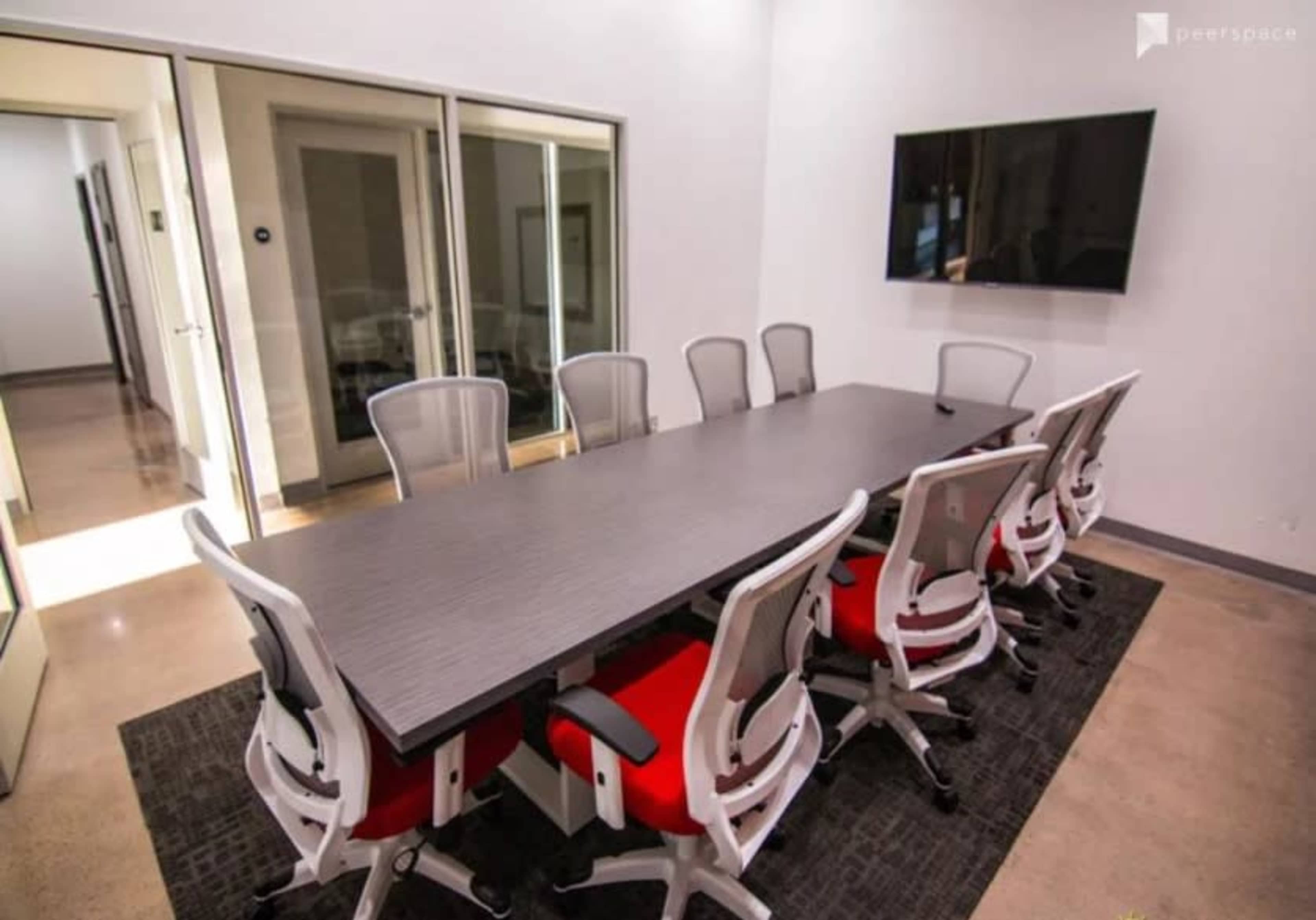 A long conference table with eight chairs is set up in a modern meeting room featuring a television display and large glass doors.
