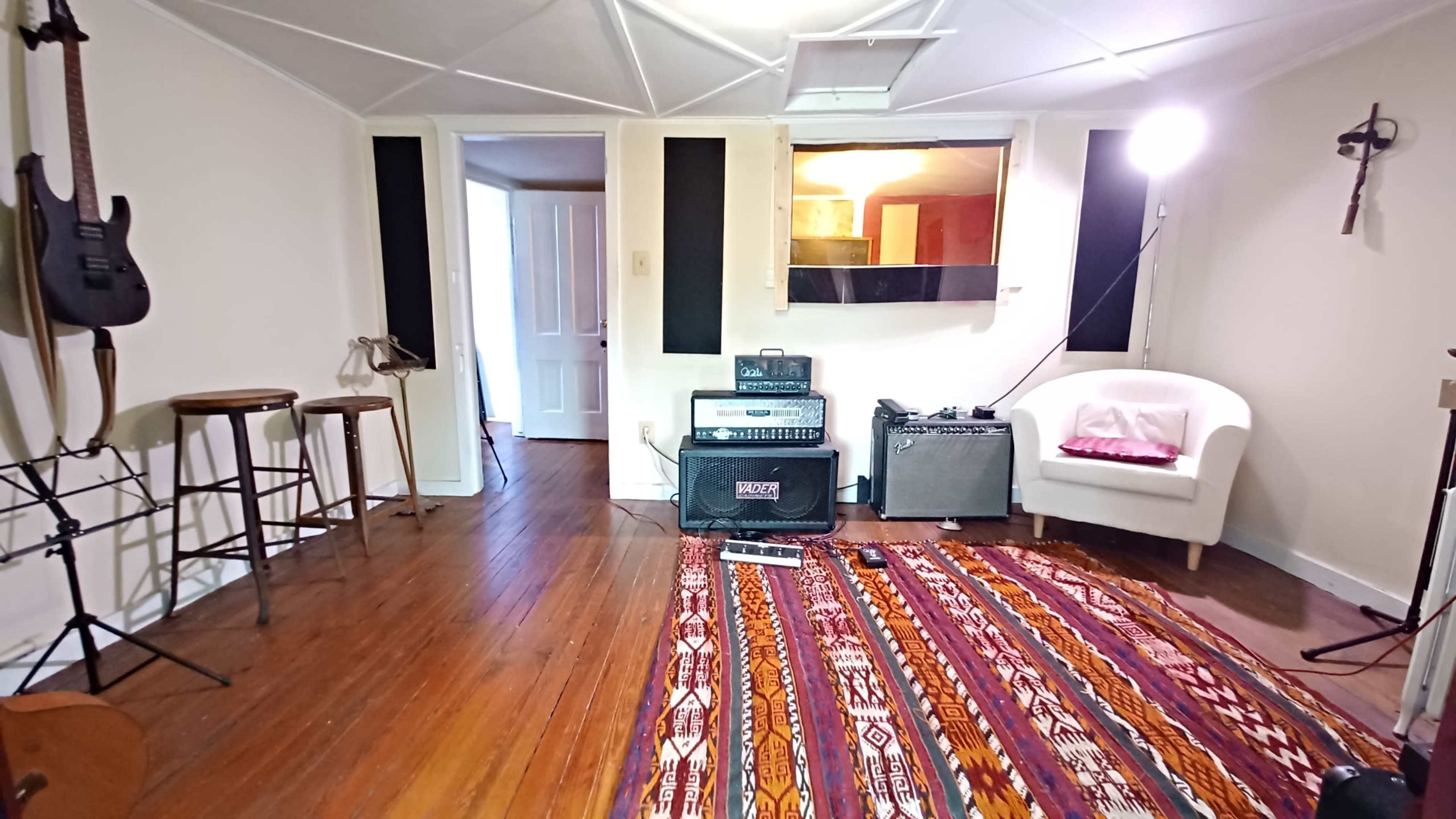 The image shows a small music studio with wooden floors, a patterned rug, an amplifier, a guitar stand, and a white chair in a corner.