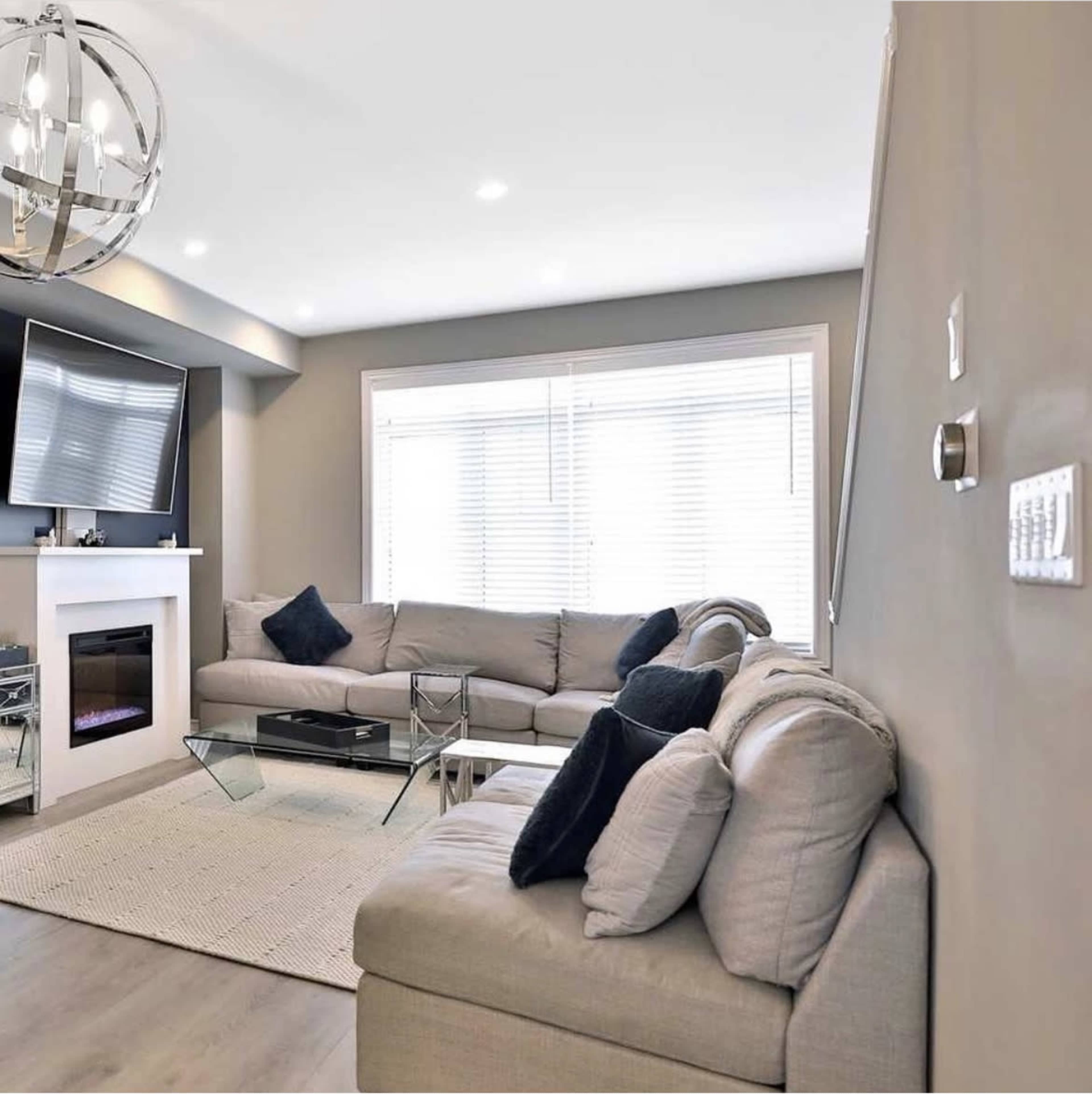 The image shows a modern living room with a gray sectional sofa, a coffee table, a television on the wall, and large windows with white blinds.