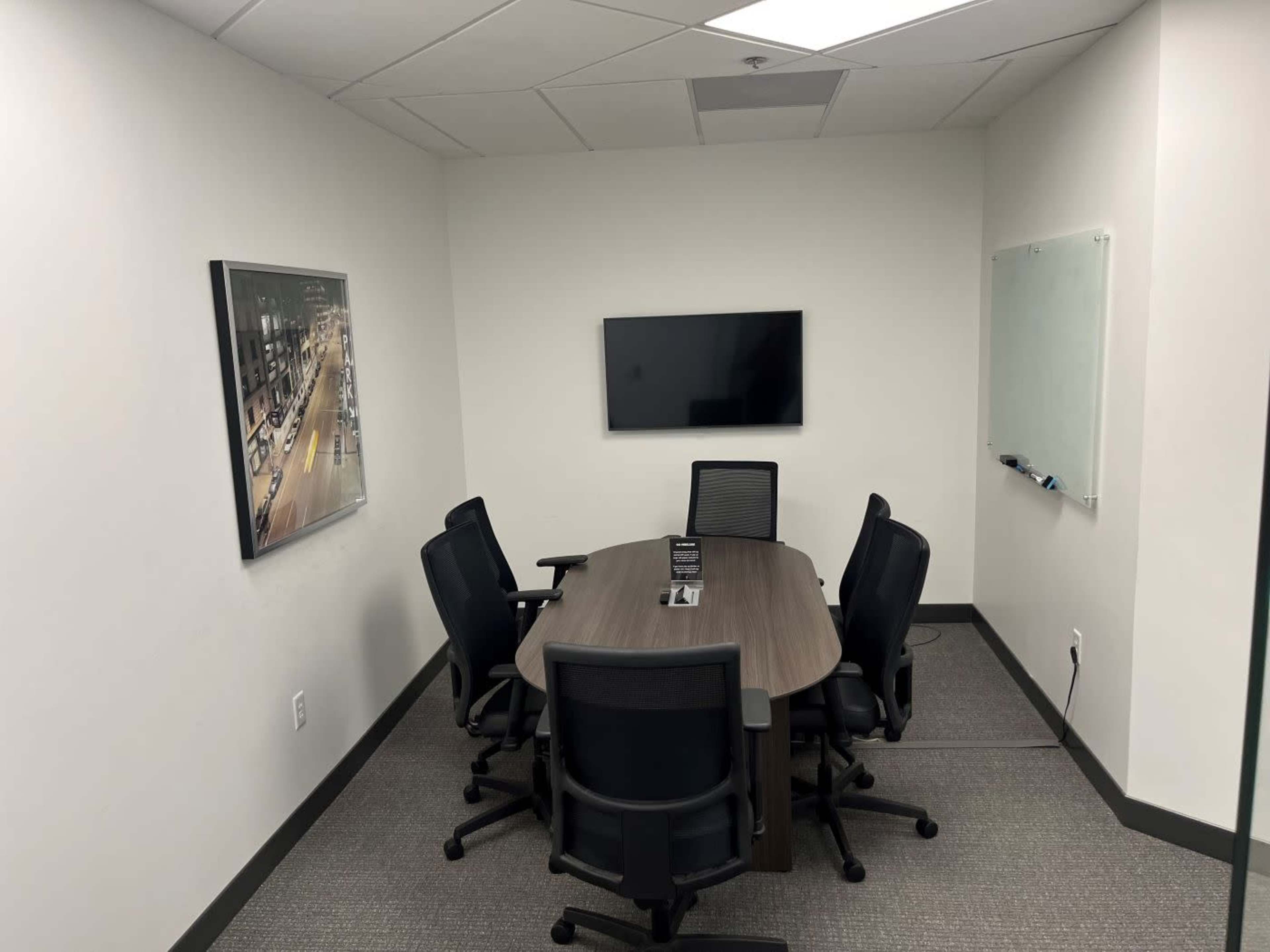 The image shows a modern conference room featuring a large table surrounded by six black chairs, a wall-mounted television, and a whiteboard.