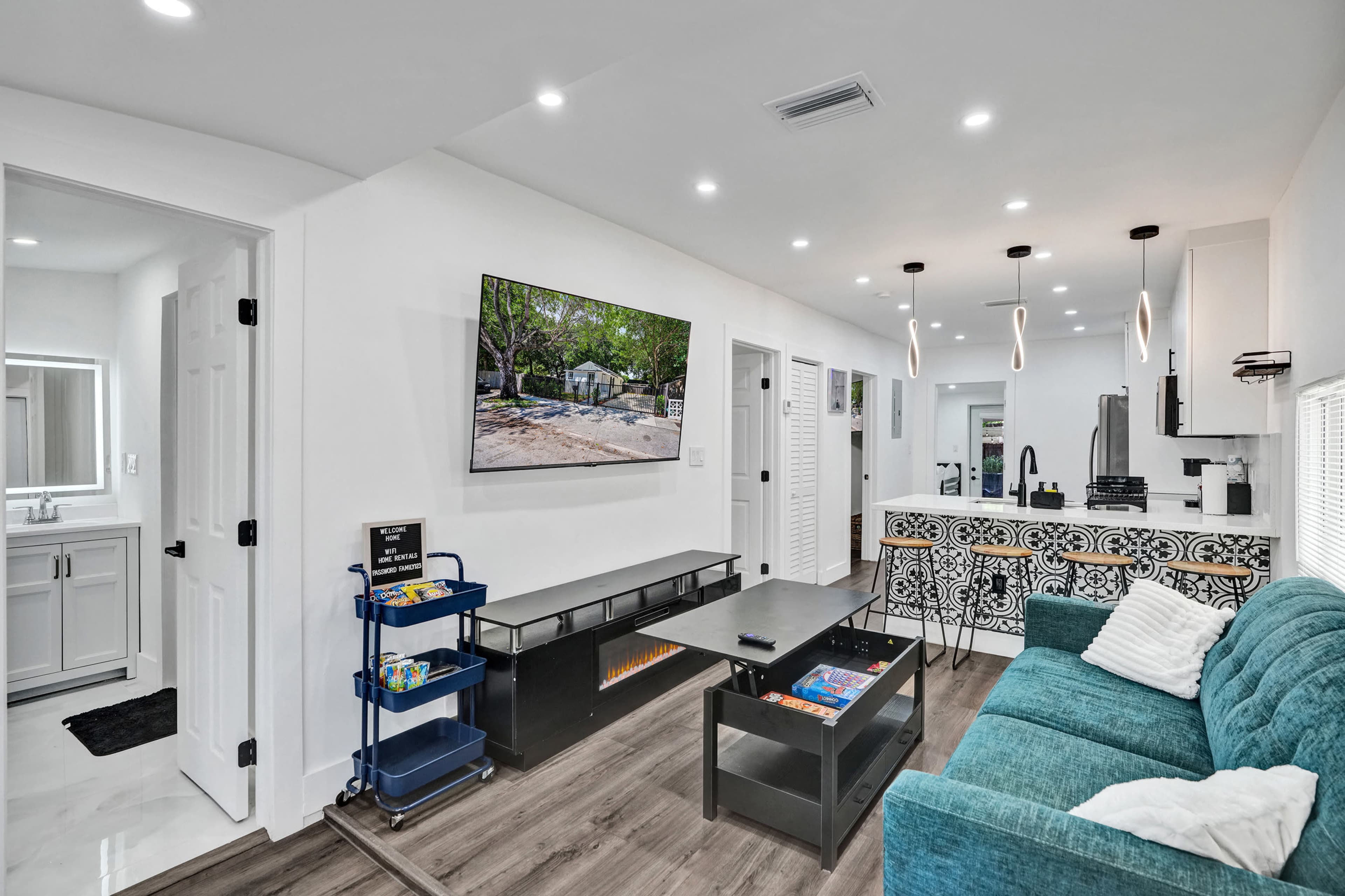 The image shows a modern living space with a turquoise couch, a wall-mounted TV, and a small kitchen area featuring decorative tiles and a cart with books.