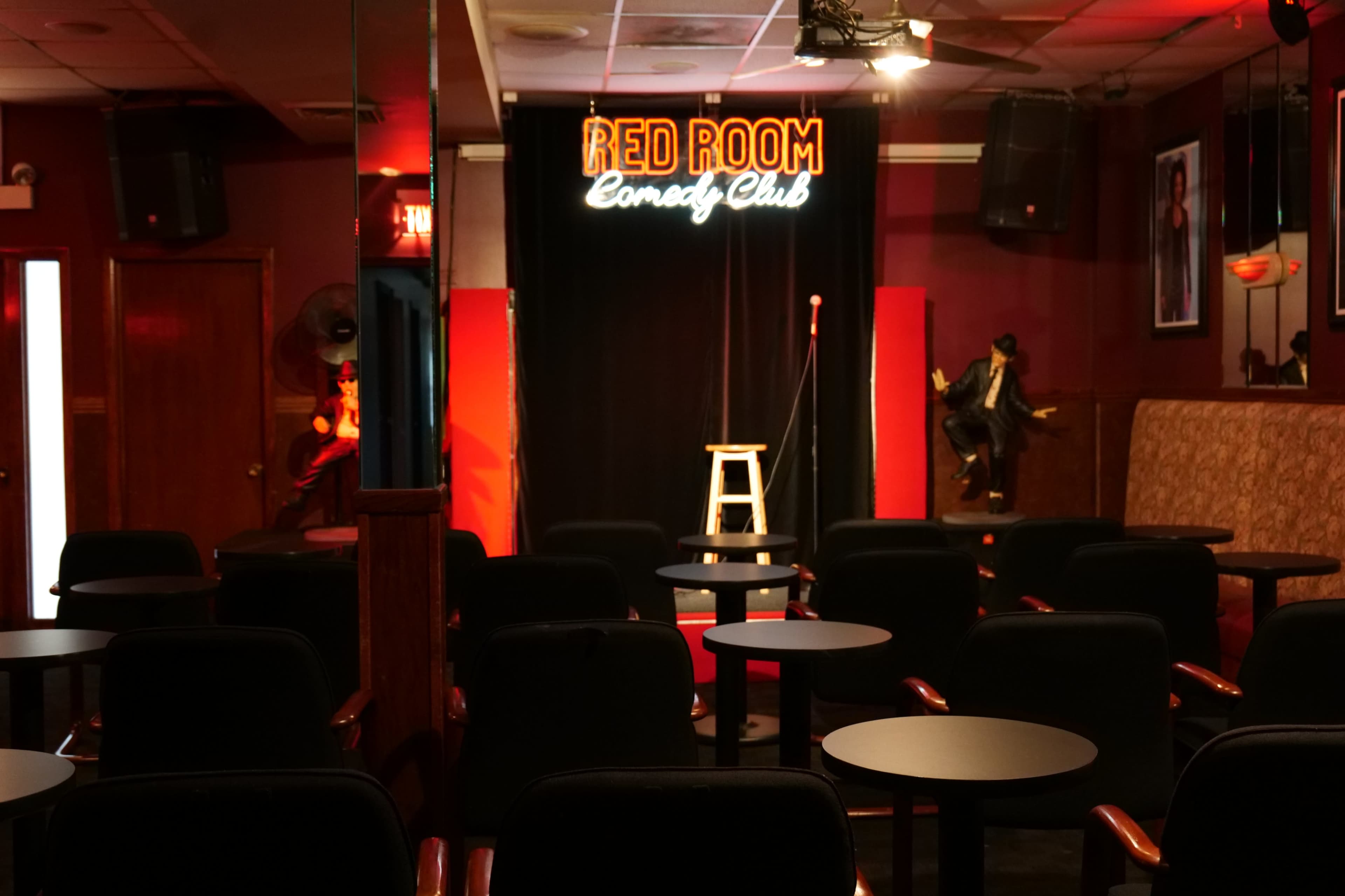 An empty comedy club with a stage featuring a microphone and a stool, illuminated by a neon sign that reads "RED ROOM Comedy Club."