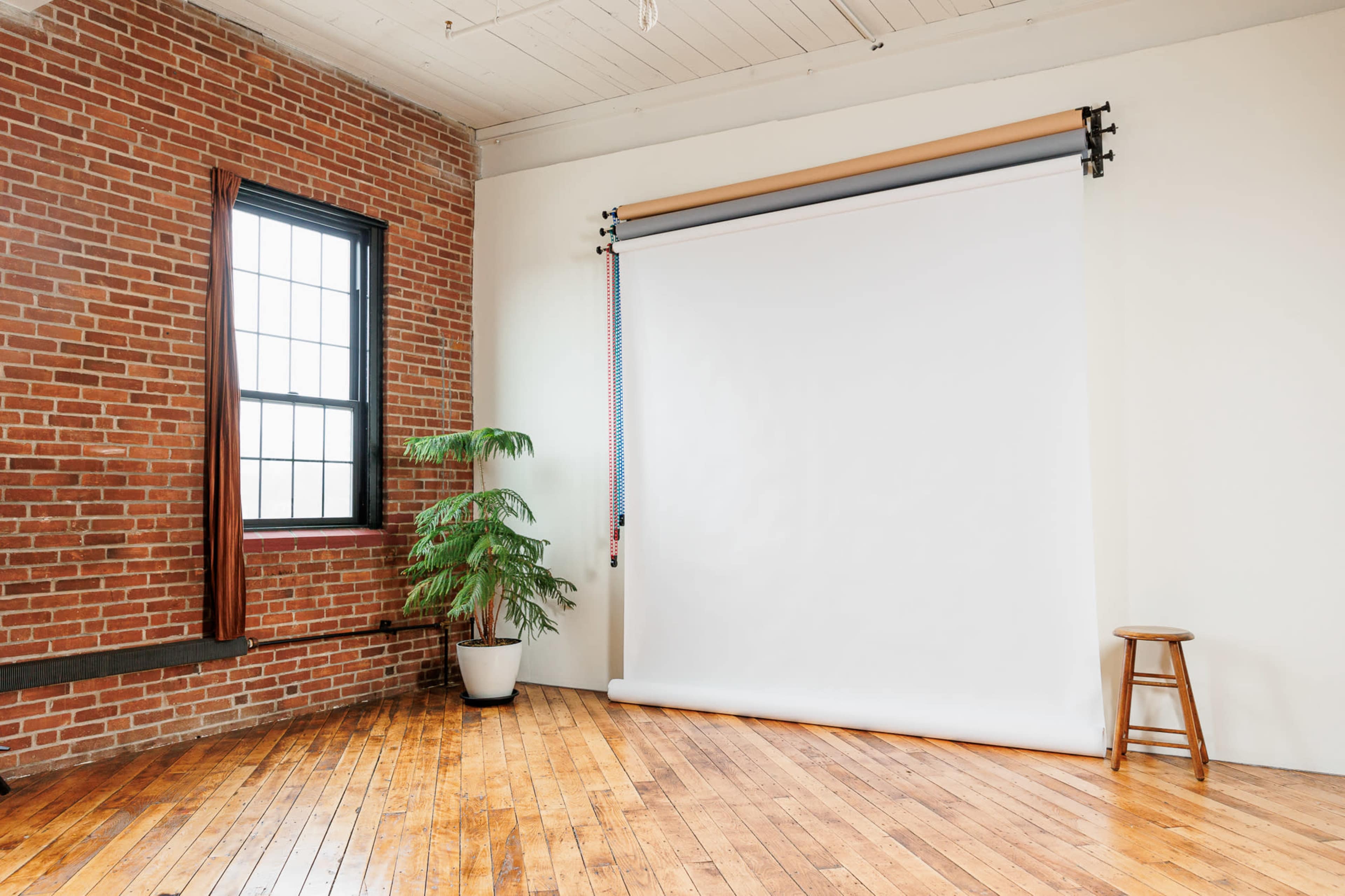 A spacious room with exposed brick walls, a white backdrop on rollers, a potted plant, and a wooden stool on a hardwood floor.