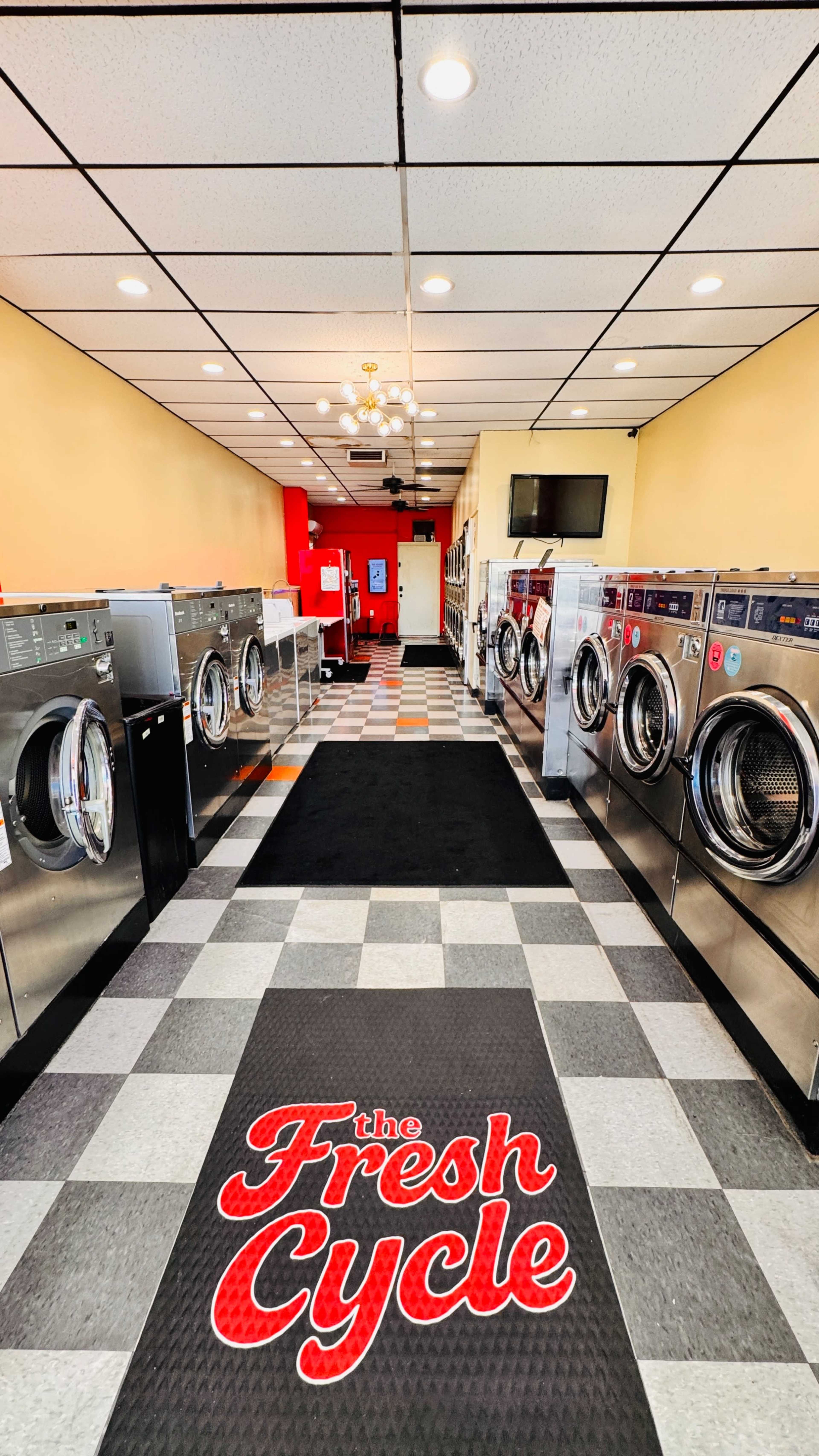 The image shows a row of commercial washing machines in a laundry facility named "The Fresh Cycle" with a checkered floor and a bright red accent wall.
