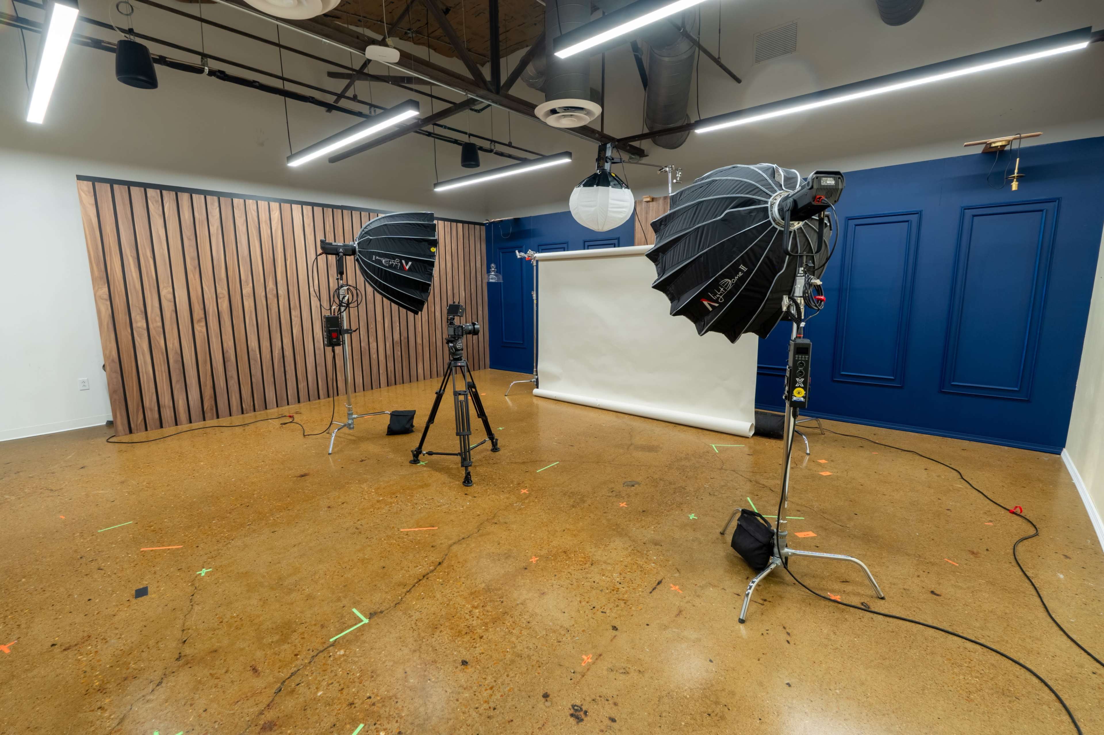 The image shows a photography studio with two large softbox lights, a camera on a tripod, and a seamless white backdrop positioned against a wall with wooden paneling.