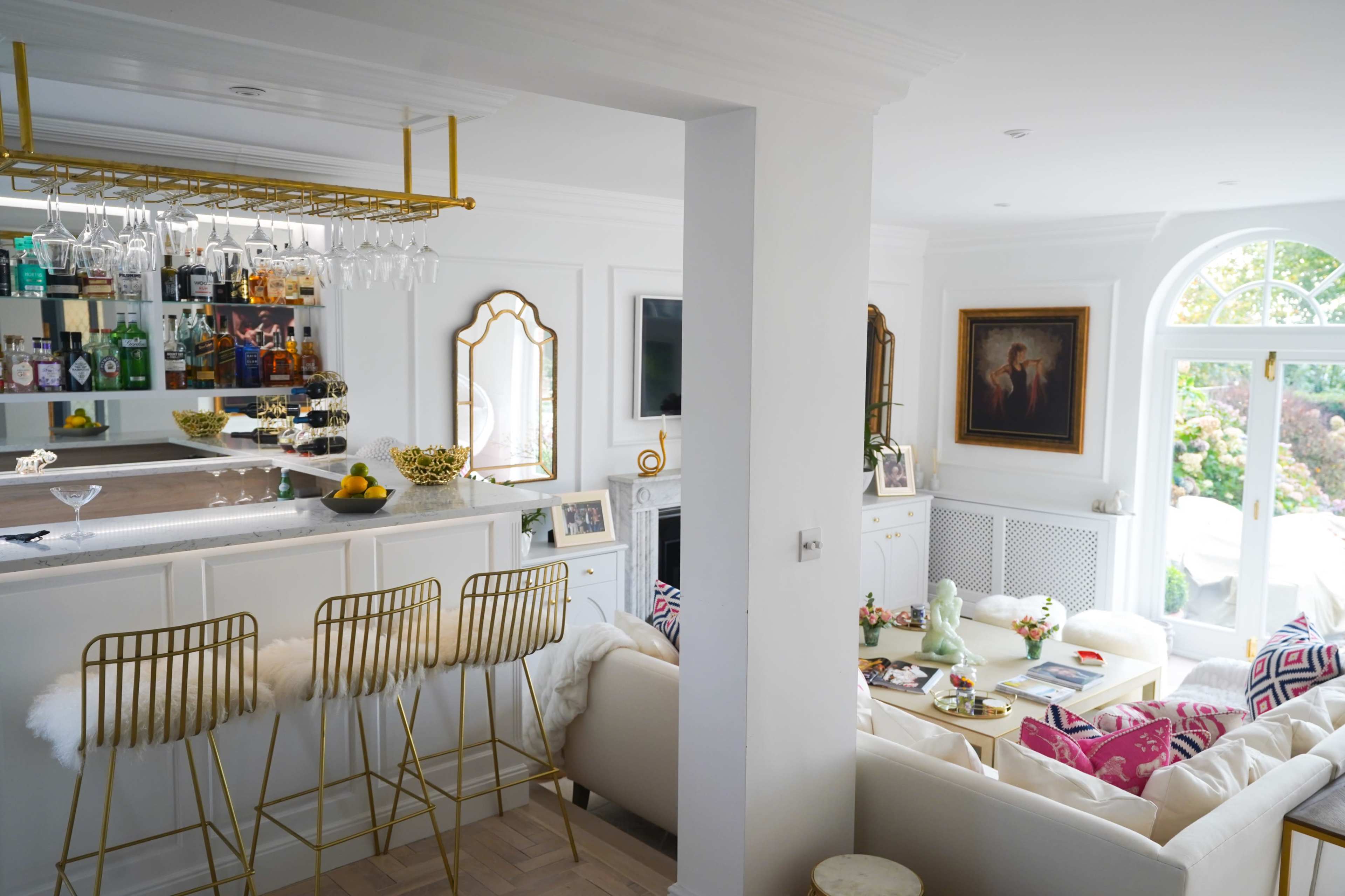 A modern living space featuring a bar area with a gold rack and several bottles, a white sofa adorned with colorful cushions, and large windows providing natural light.