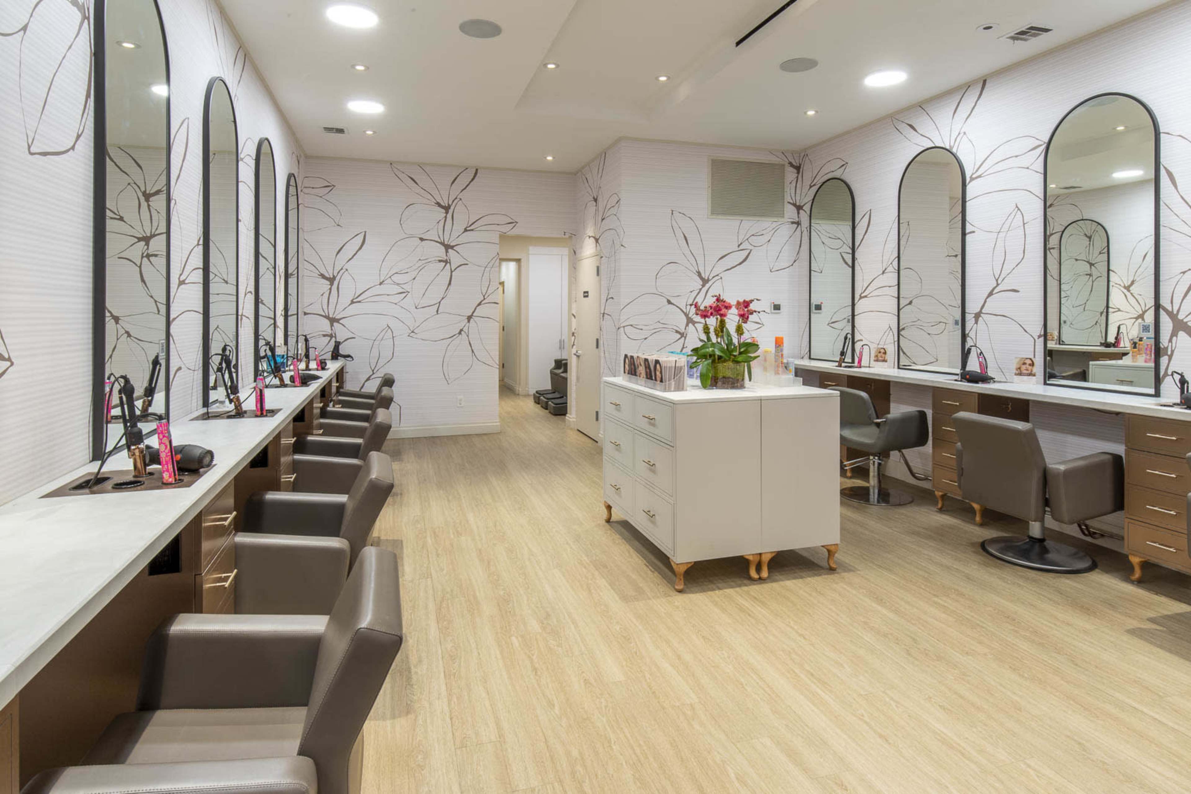 The image shows a beauty salon interior with multiple styling stations, mirrors, and a central storage unit, all featuring a floral wallpaper design.