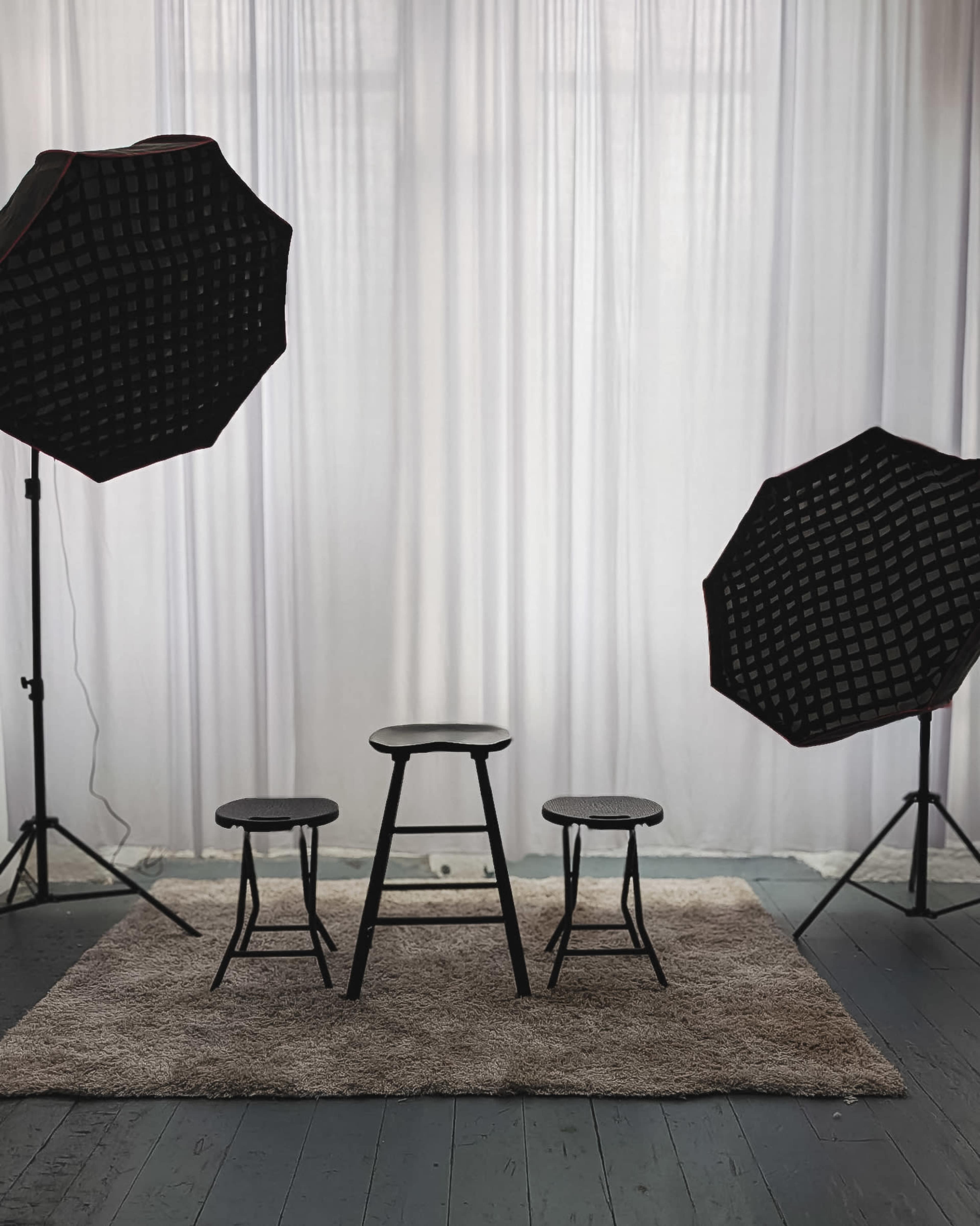 The image shows a photography setup with three black stools arranged on a rug, flanked by two large softboxes directed towards a white curtain backdrop.