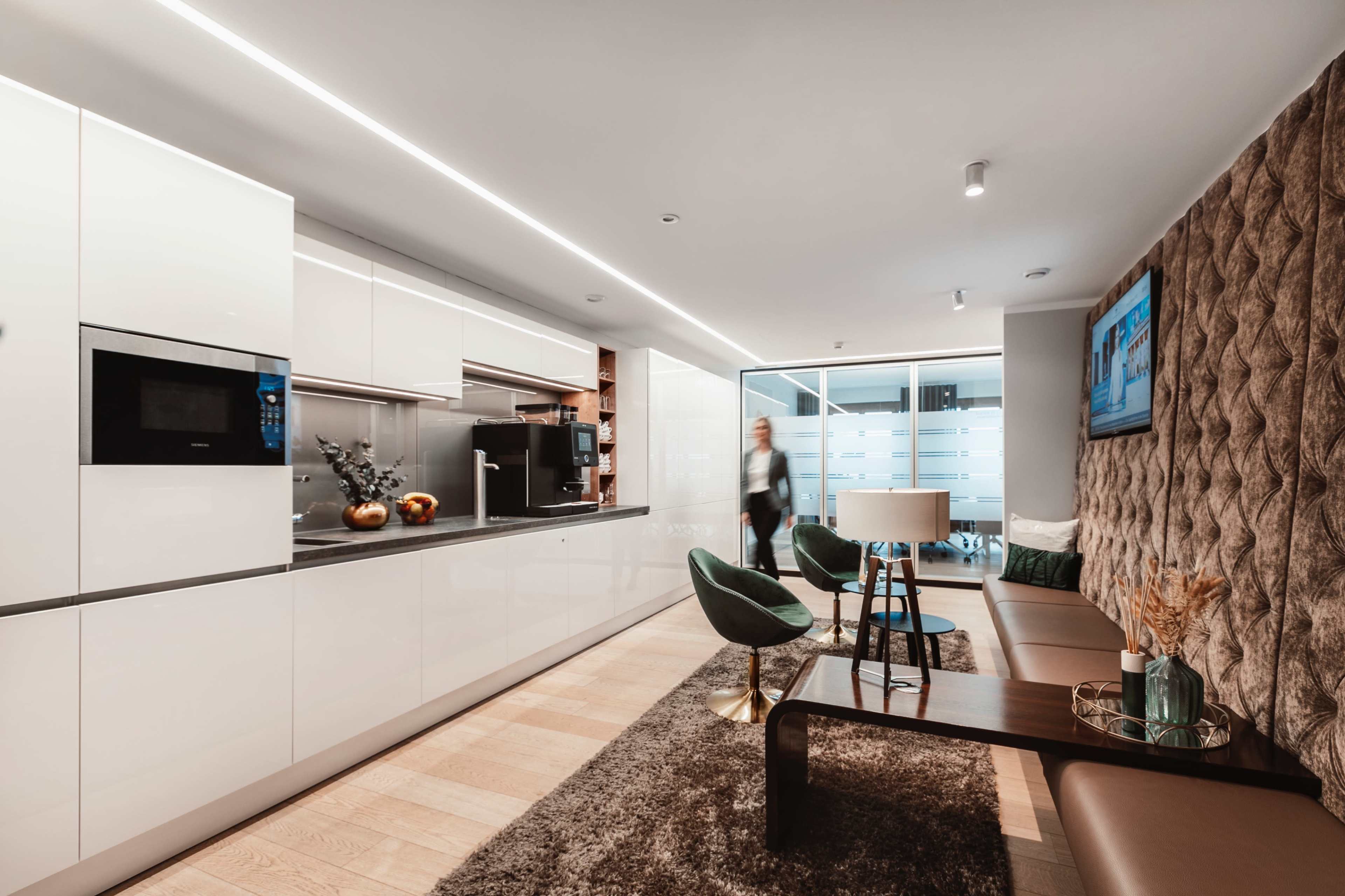 A modern kitchen and living area feature sleek white cabinets, a brown tufted wall, and green chairs, with a table and decor items.