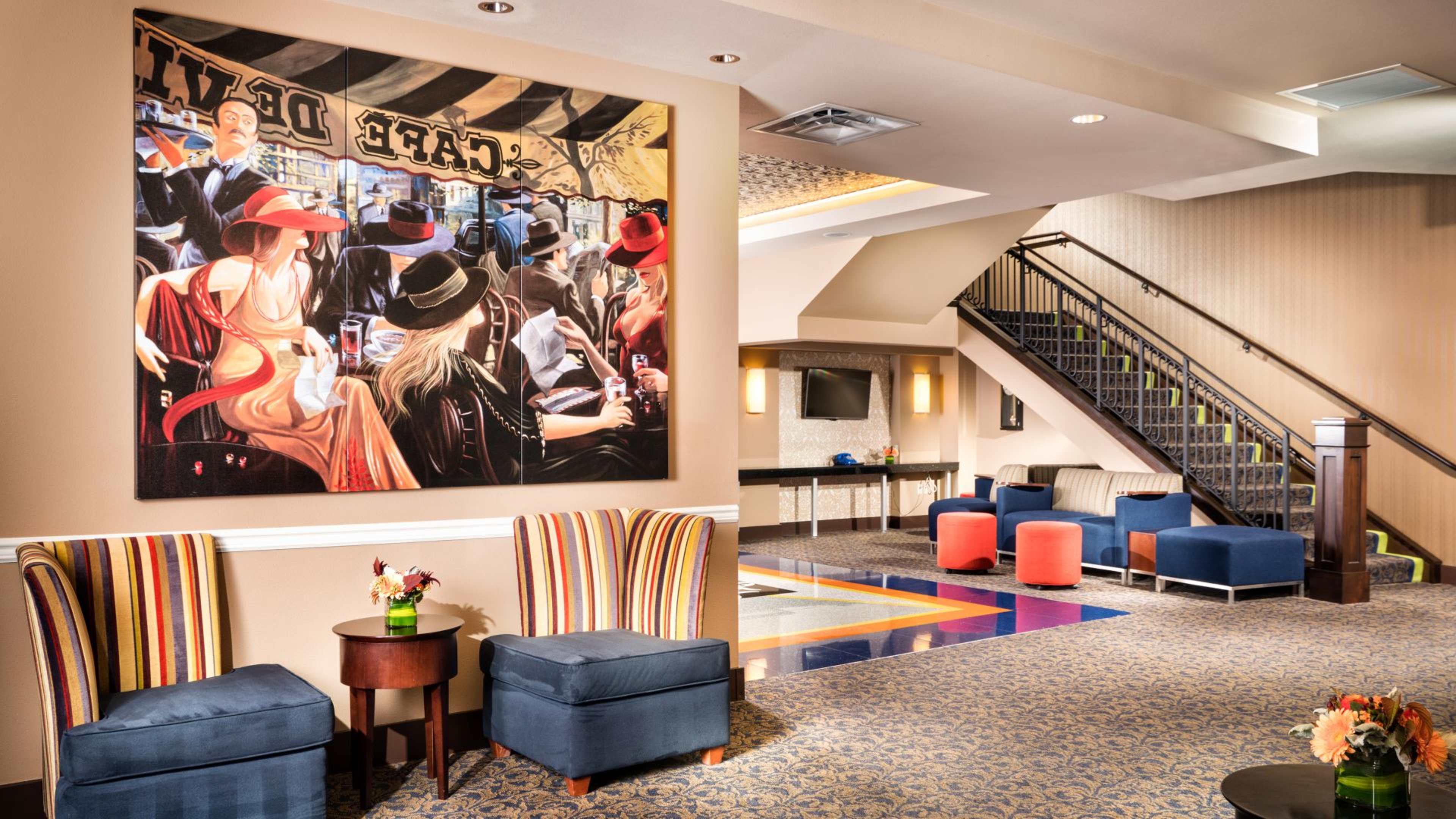 A hotel lobby featuring two striped chairs, a small table with flowers, a large painting on the wall, and a staircase leading to an upper level.