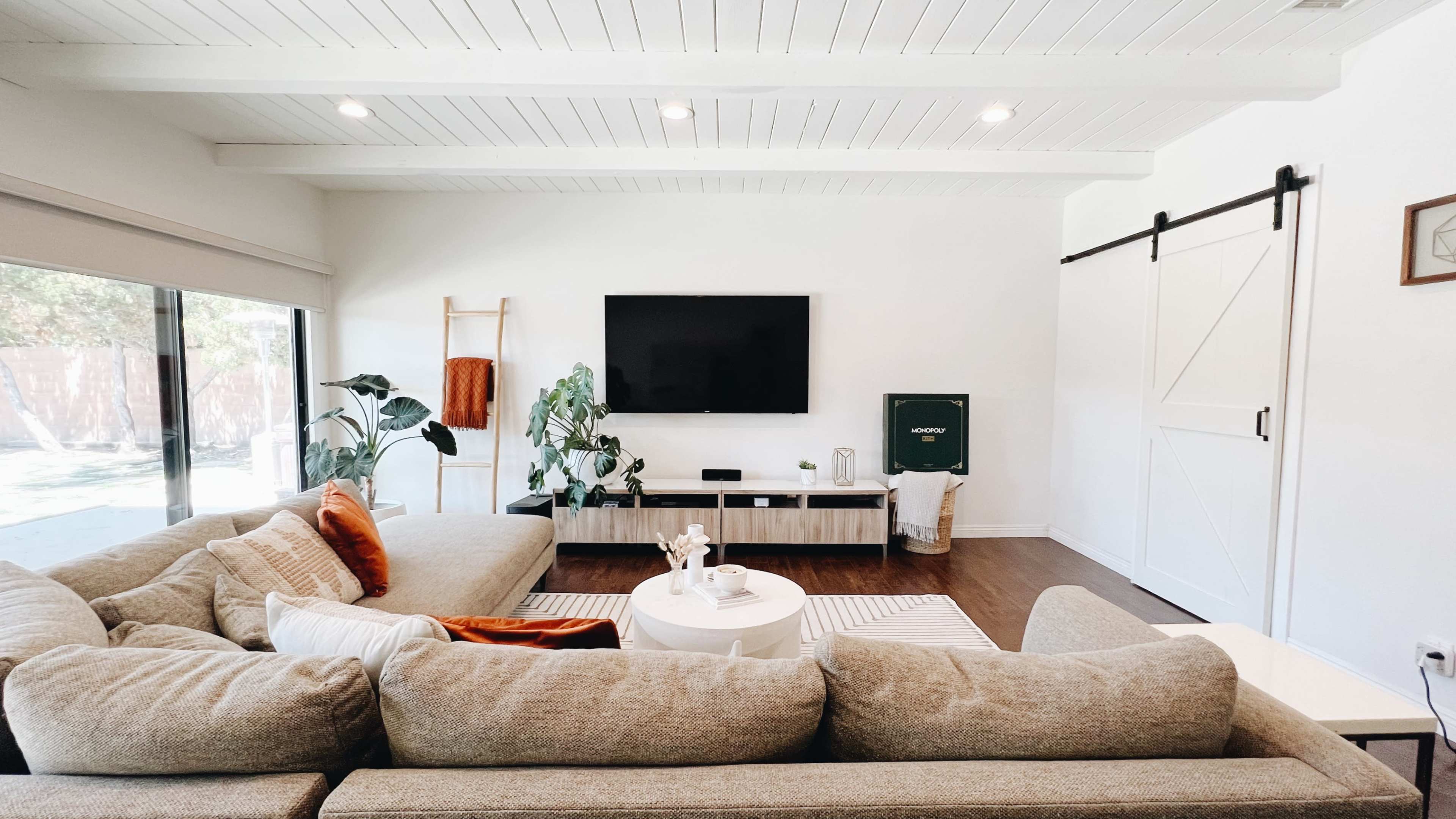 A modern living room features a large sectional sofa, a wall-mounted television, and a light wood media console with plants and decor.
