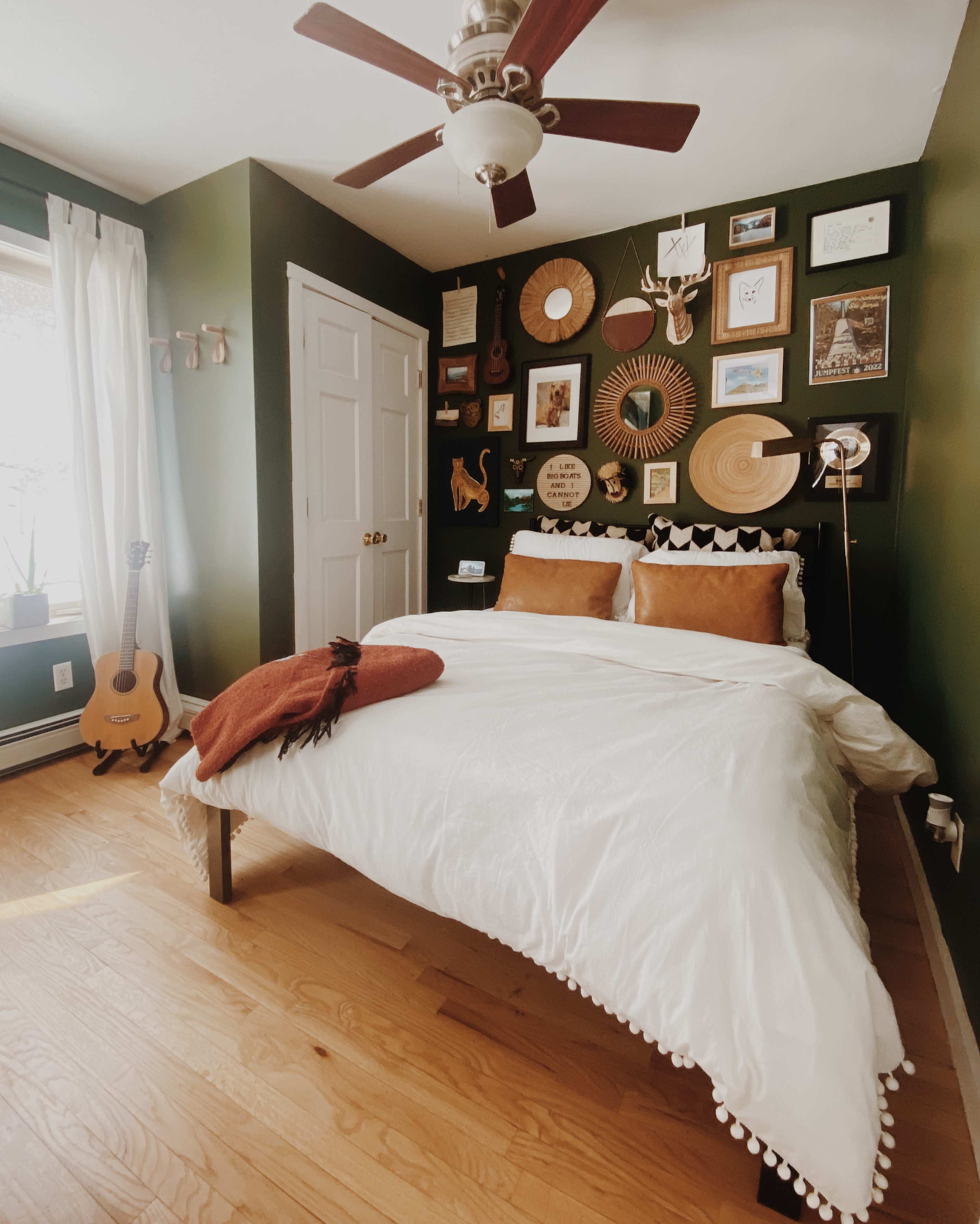 A bed with white bedding and two brown pillows is positioned against a dark green wall adorned with various framed artworks, complemented by a wooden floor and a small guitar in the corner.