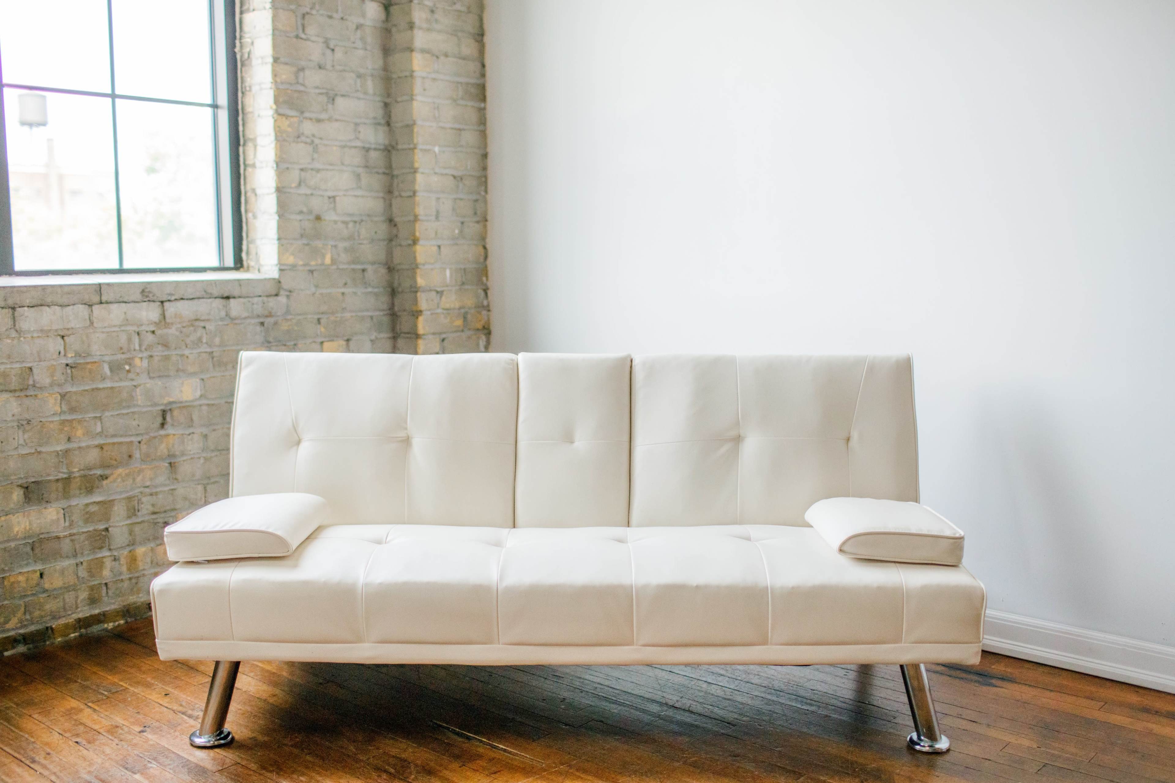 A light-colored sofa with two armrests is positioned against a wall in a room with a large window and exposed brick.