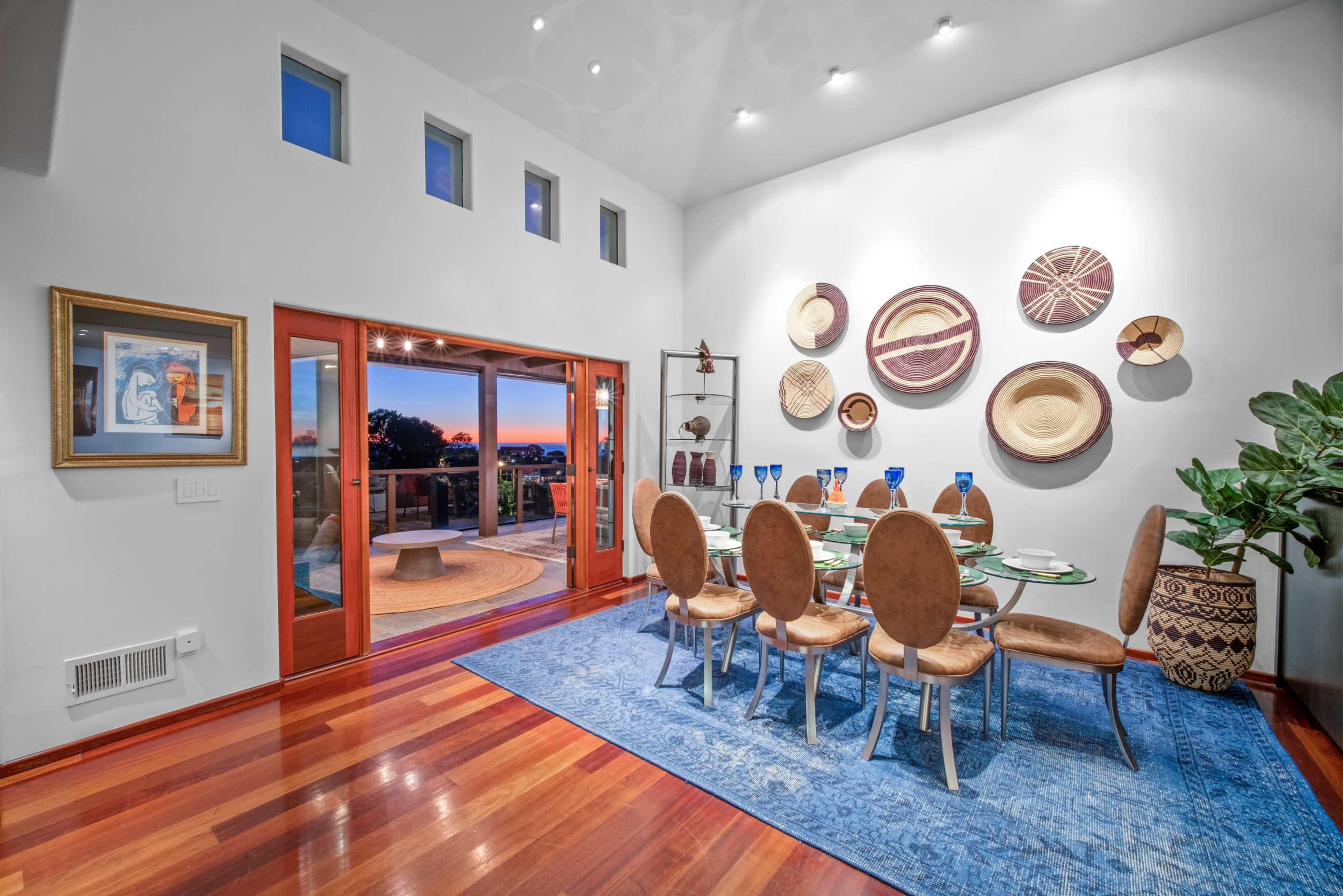 A modern dining room with a glass table set for eight, surrounded by brown chairs, against a backdrop of colorful woven wall art and large windows opening to a scenic view.