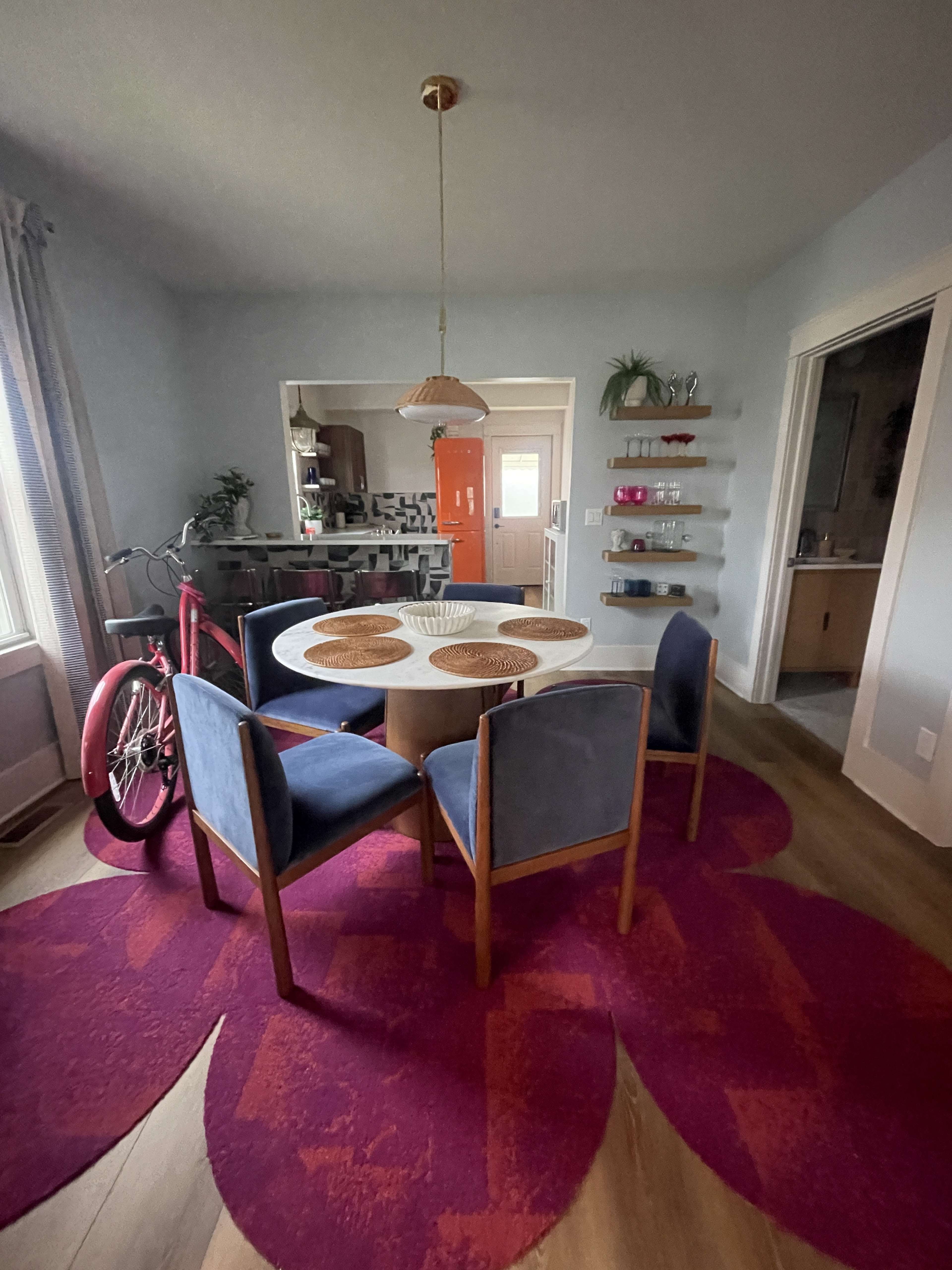 A dining area with a round table surrounded by four chairs, a bicycle parked nearby, and colorful decor on the walls.