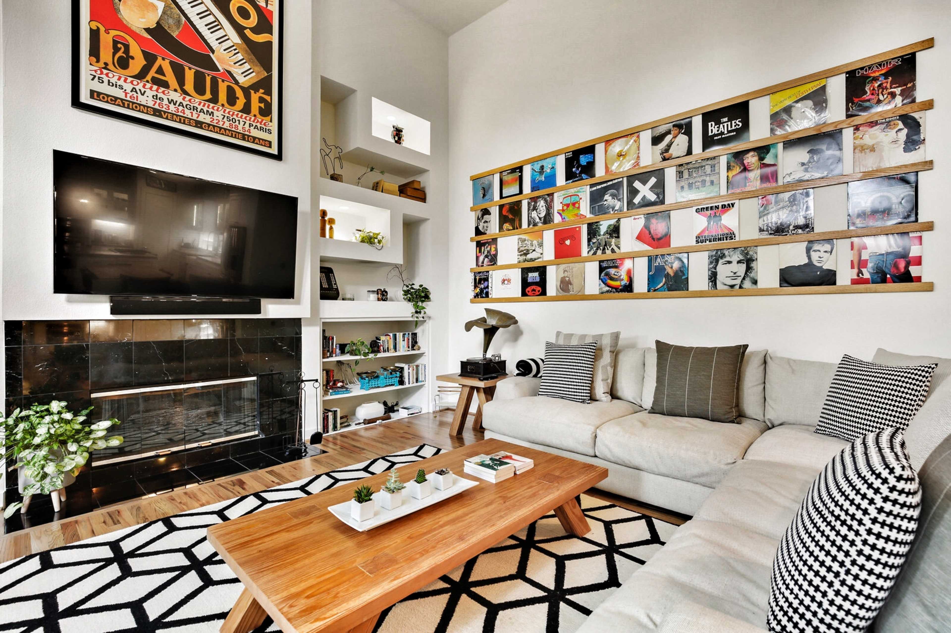 A light-filled living room features a beige couch, a wooden coffee table, and a wall displaying a collection of framed album covers alongside a mounted flat-screen TV.