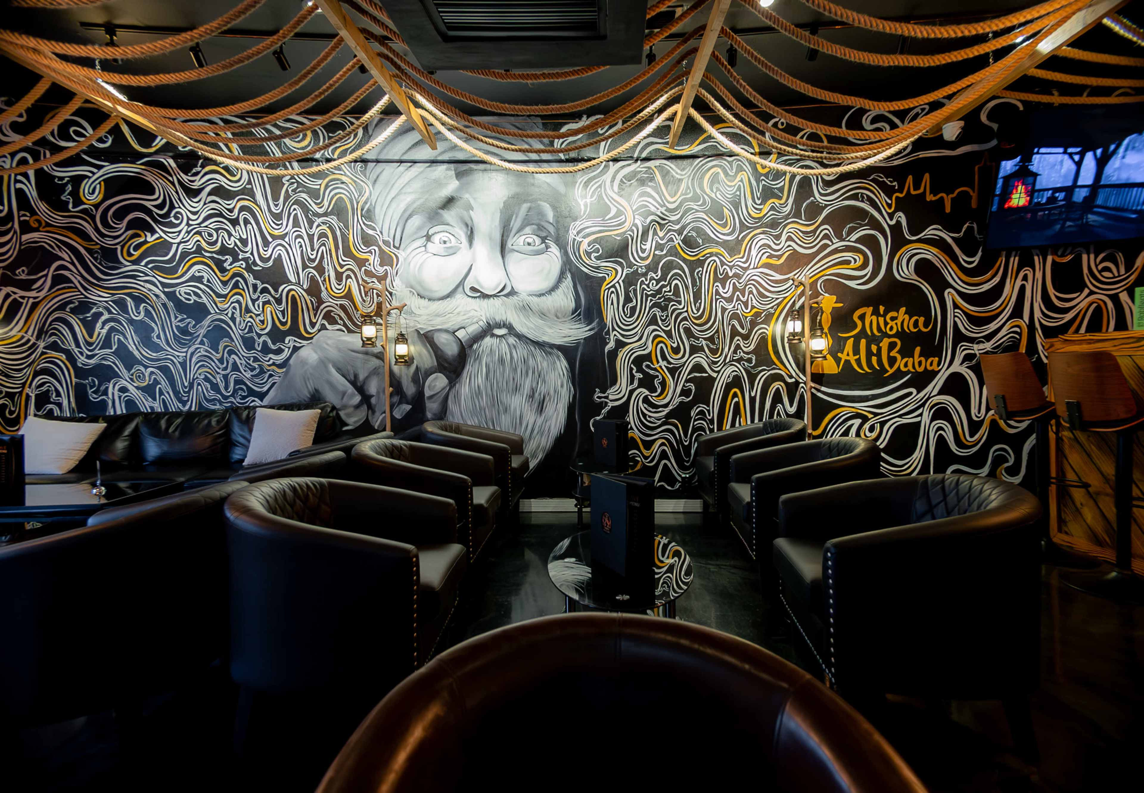 A stylish lounge area featuring black walls adorned with a large mural of a bearded man, surrounded by decorative rope elements and modern seating arrangements.