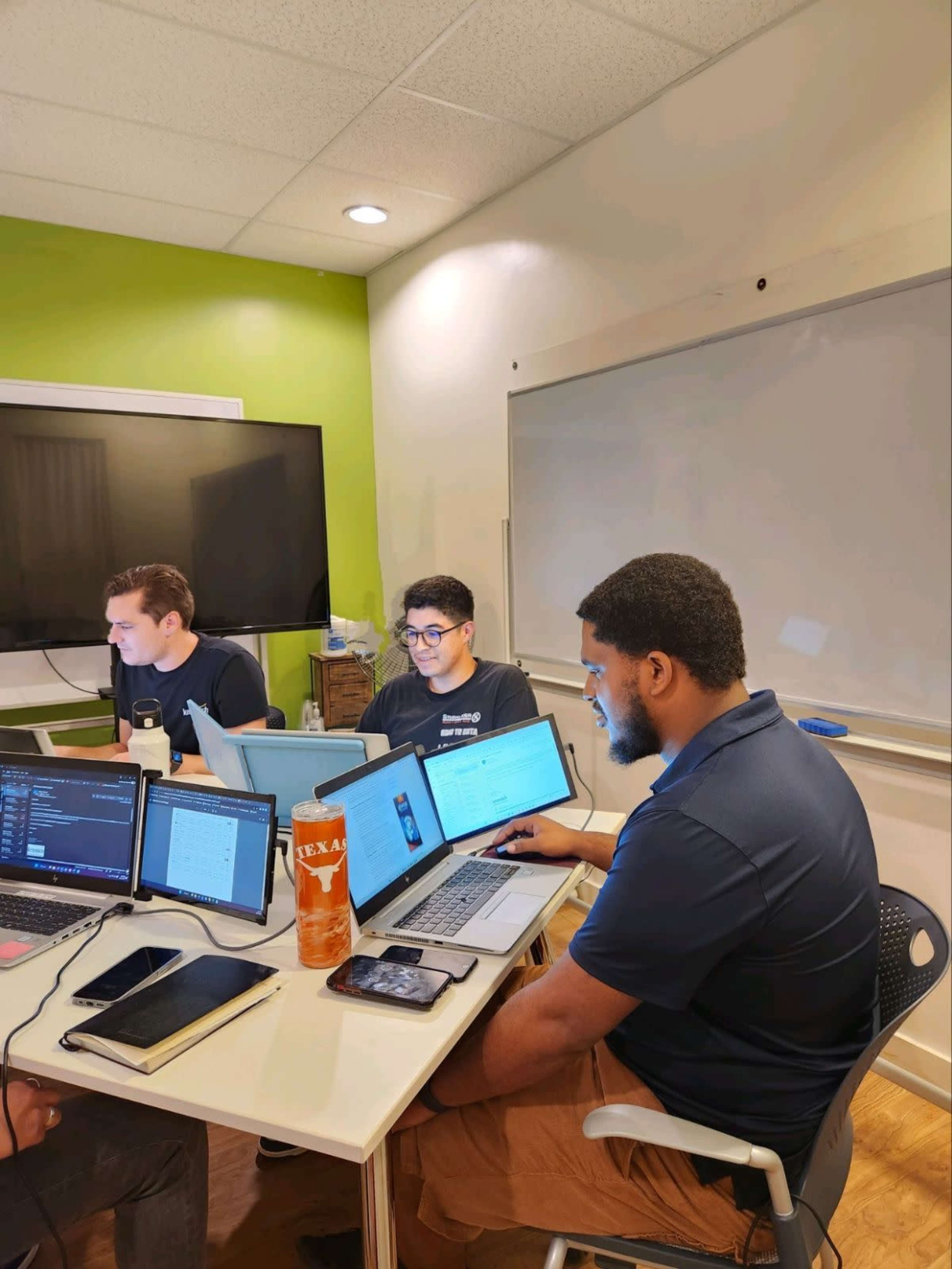 Three individuals are working at a table with laptops and notebooks in a room featuring a green accent wall and a whiteboard.