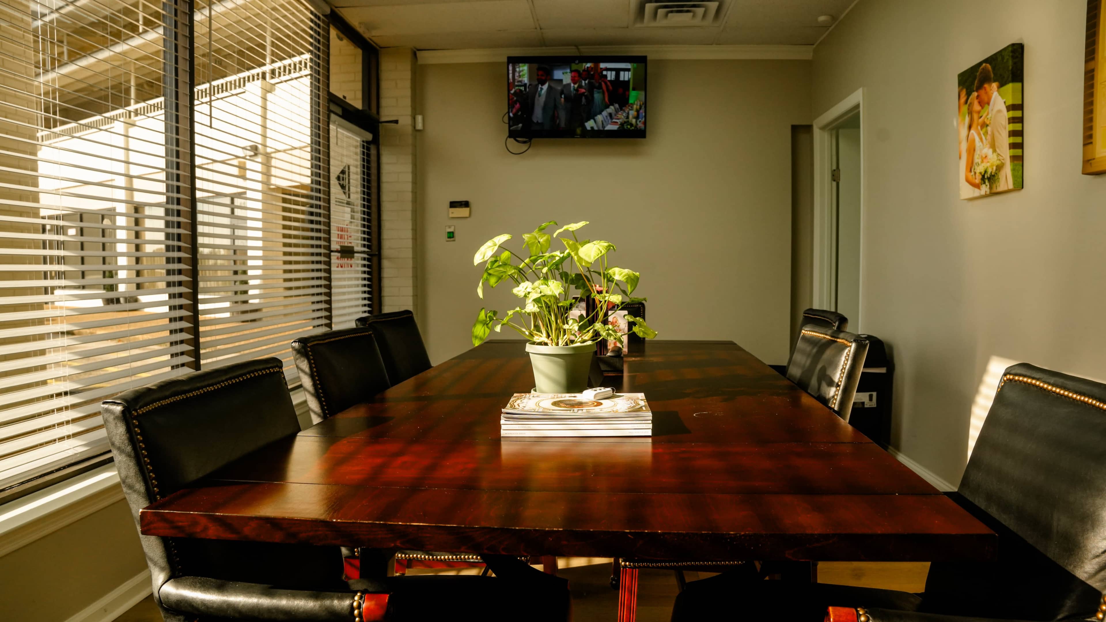 A long wooden conference table with eight black chairs is set under a television displaying a program, while a potted plant sits in the center.