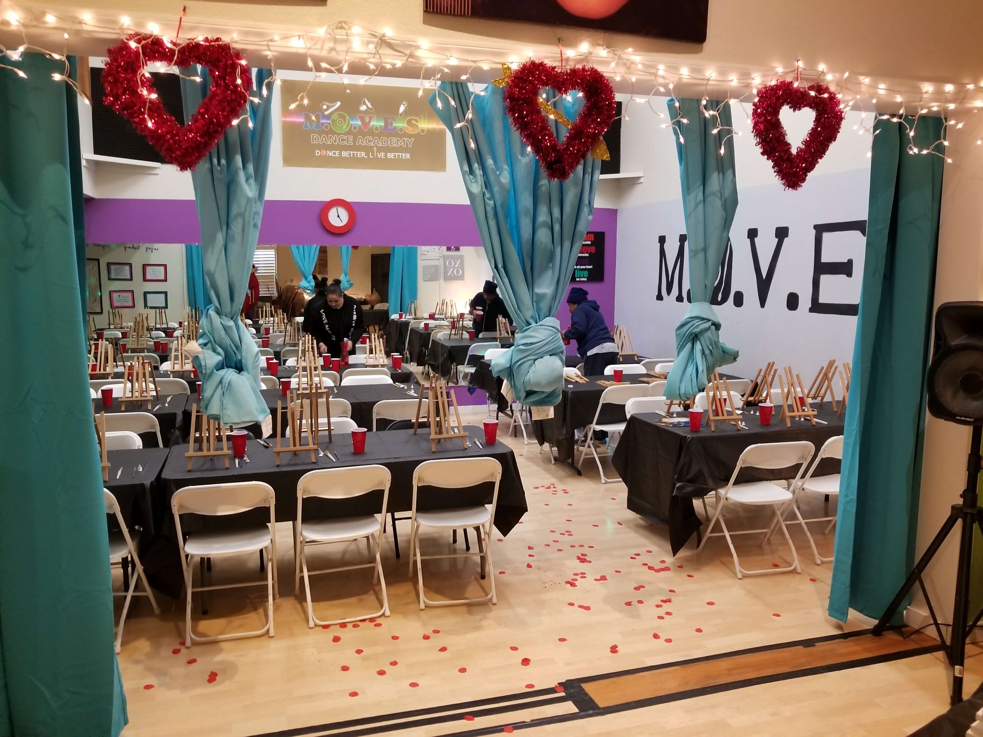 A decorated room with tables, chairs, and easels set up for an event, featuring heart-shaped decorations and rose petals scattered on the floor.