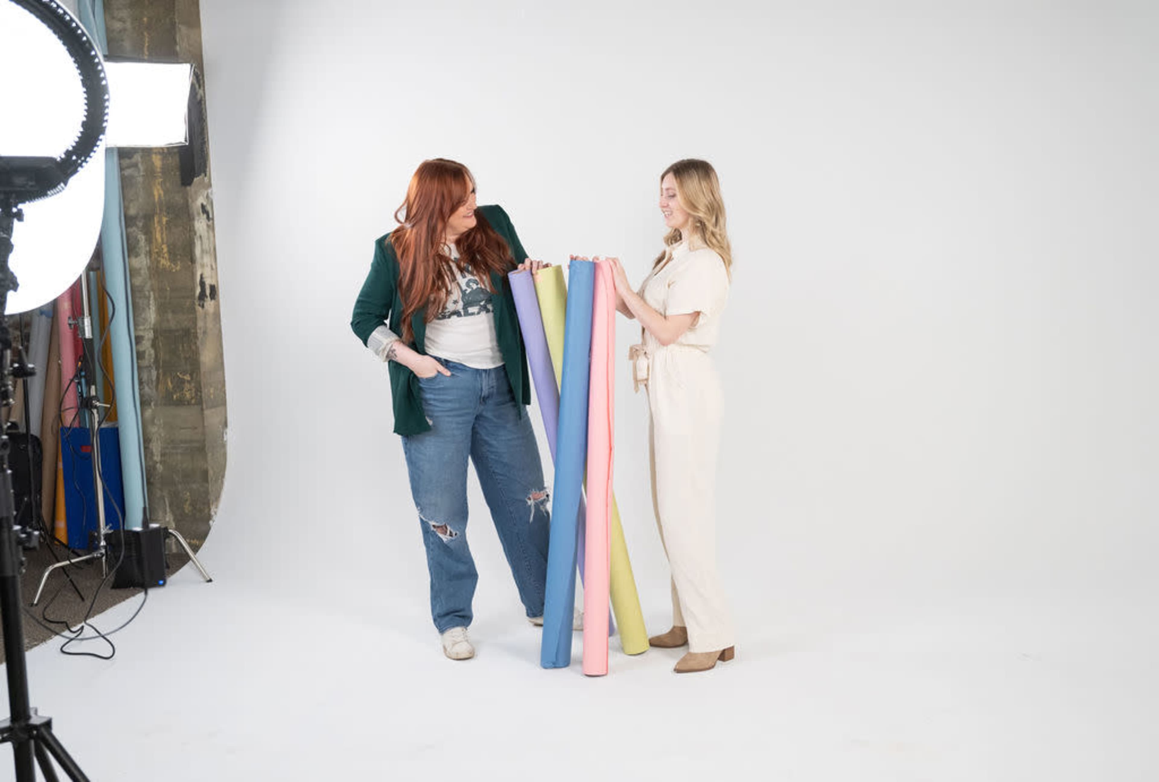 Two women stand in a studio, holding rolls of colored backdrop paper while posing next to a large light source.