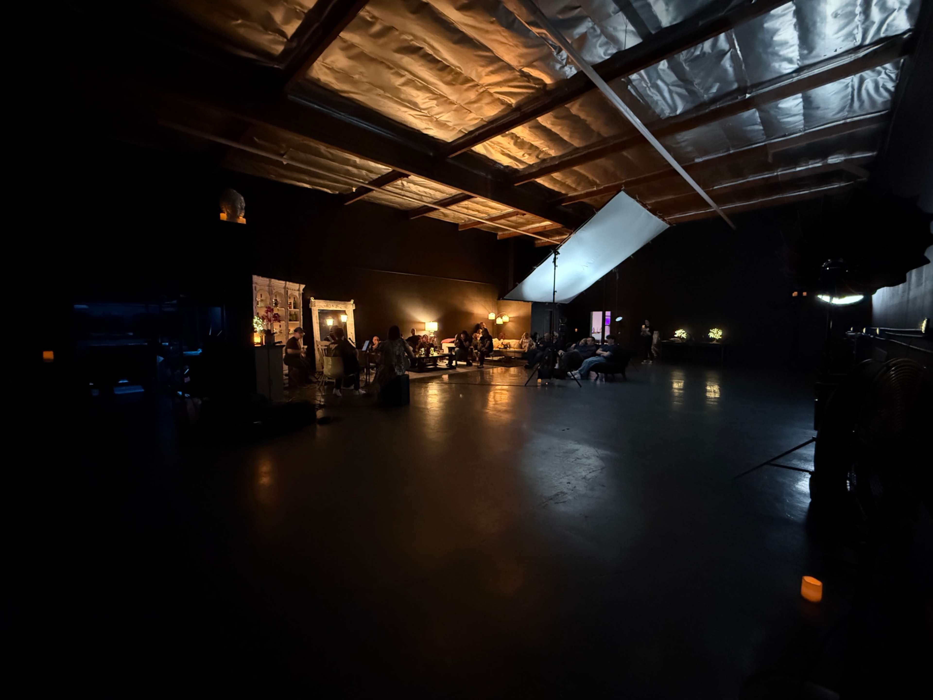 People are seated in a dimly lit studio space, with a large ceiling and various props scattered around.