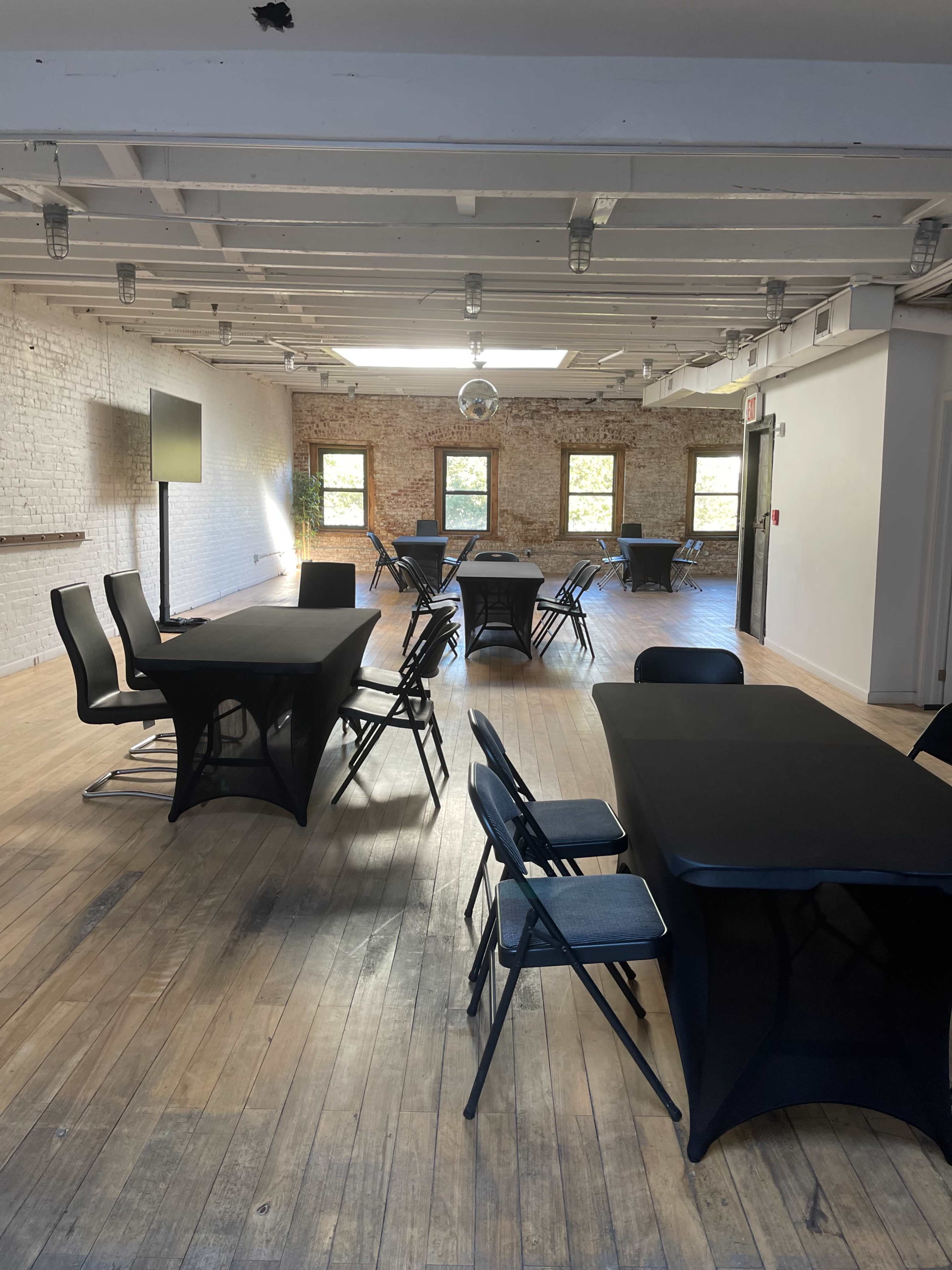 A spacious room with wooden floors, featuring several black tables and chairs arranged throughout, and large windows allowing natural light to enter.