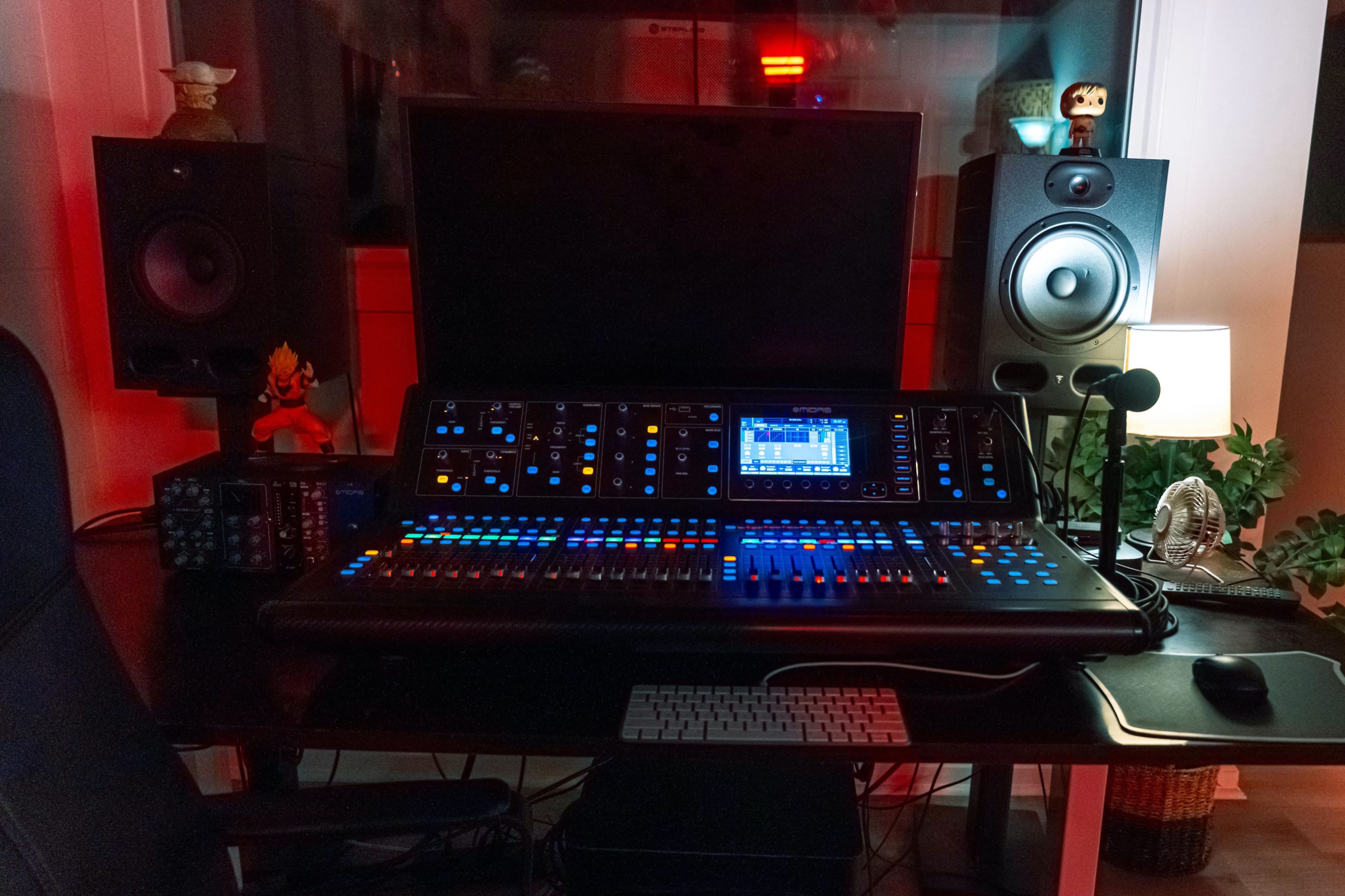 The image shows a music production workstation featuring a digital mixing console, studio monitors, a computer monitor, and a keyboard, set in a well-lit room with decorative items.