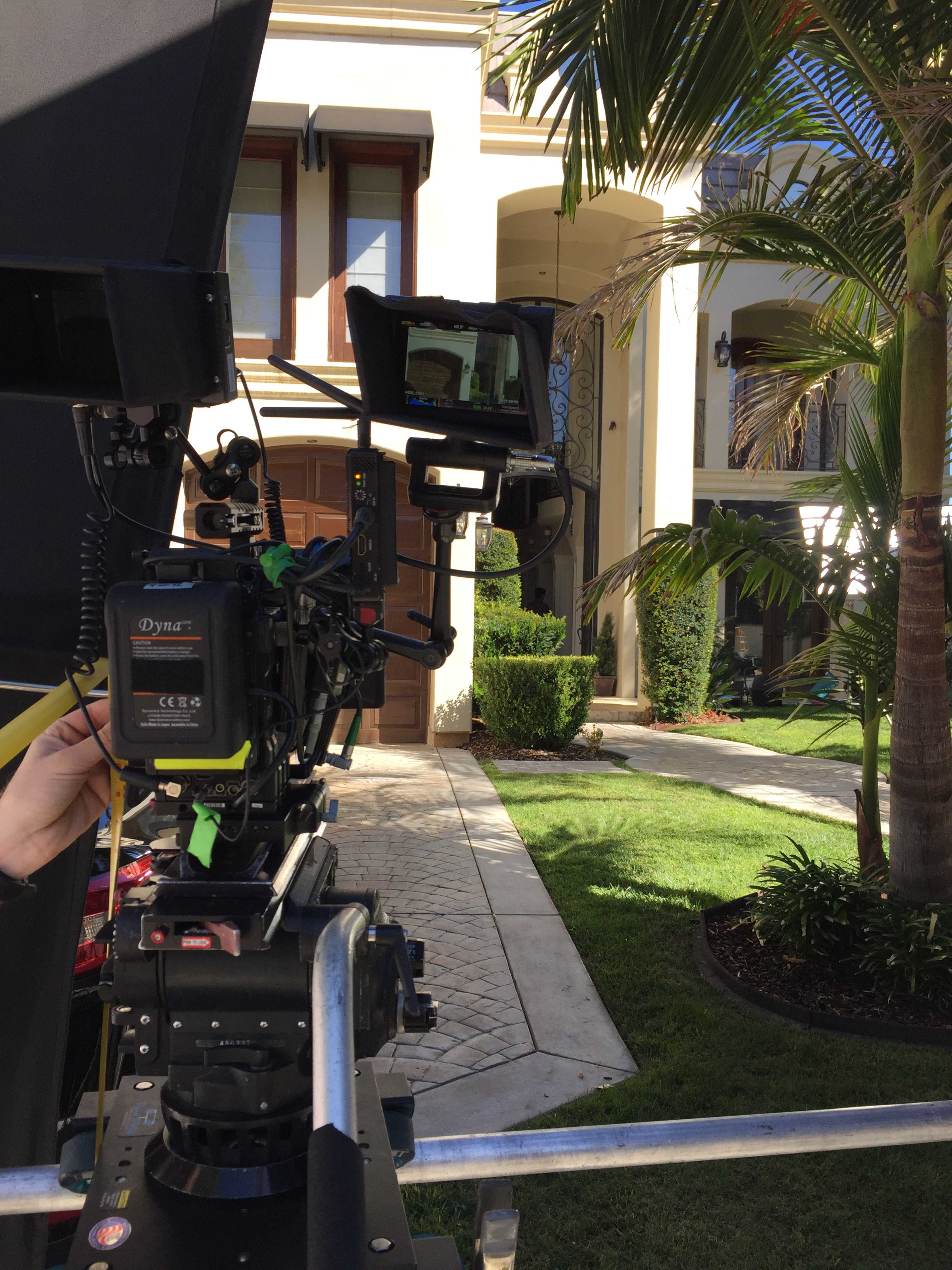 A camera setup is positioned in front of a large house, with palm trees and manicured landscaping visible in the background.