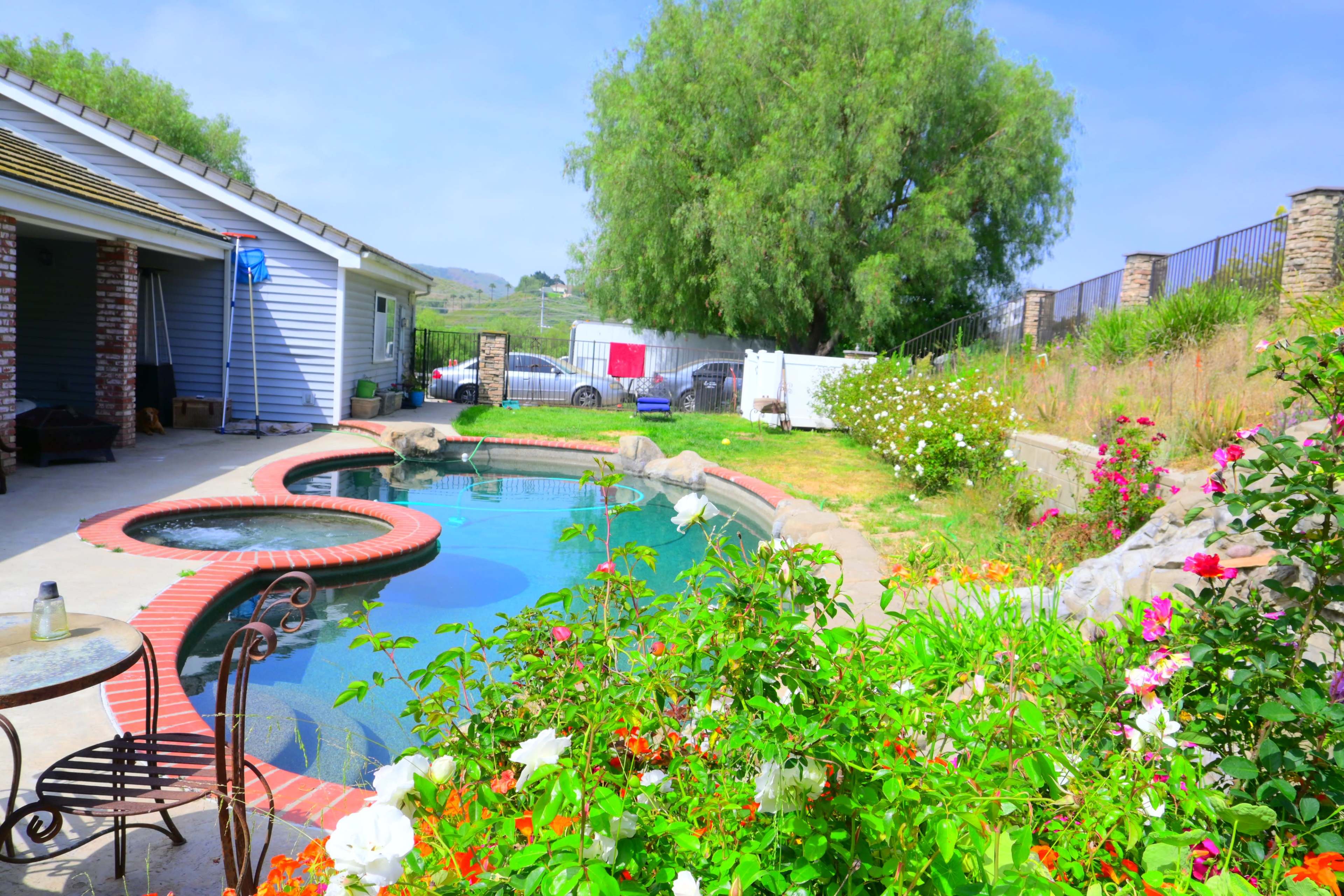 A backyard features a swimming pool with a hot tub, surrounded by colorful flowers and a grassy area, with a house visible in the background.