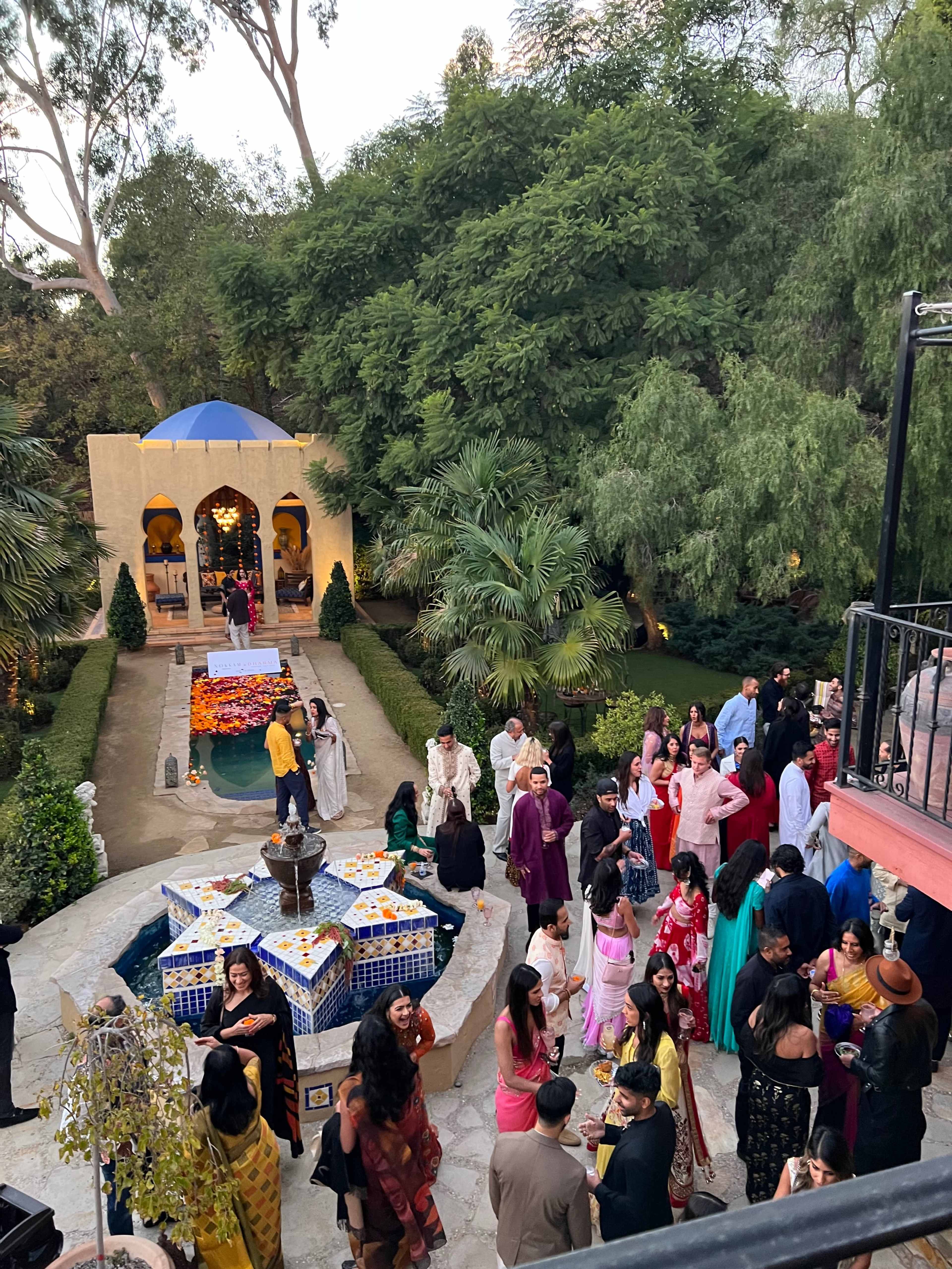 A vibrant outdoor gathering takes place in a lush garden, featuring guests in traditional attire around a decorated fountain and a table of colorful decorations.