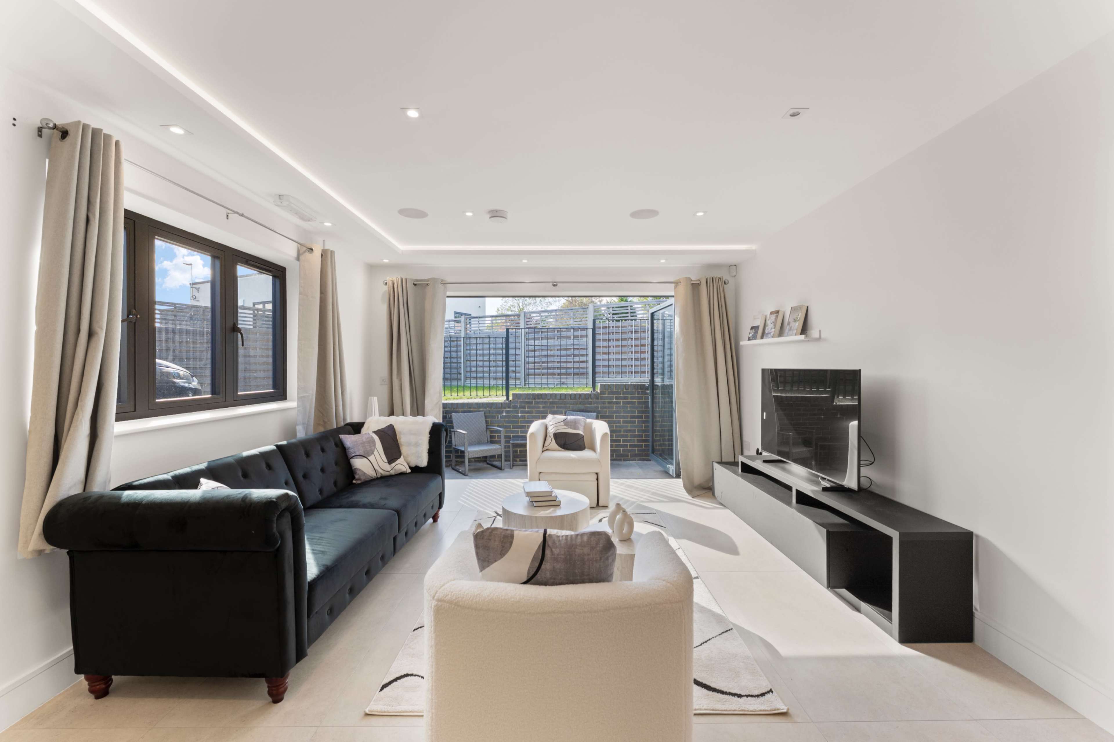 The living room features a black sofa, a white armchair, and a television mounted on a black unit, with large windows providing a view of an outdoor patio area.