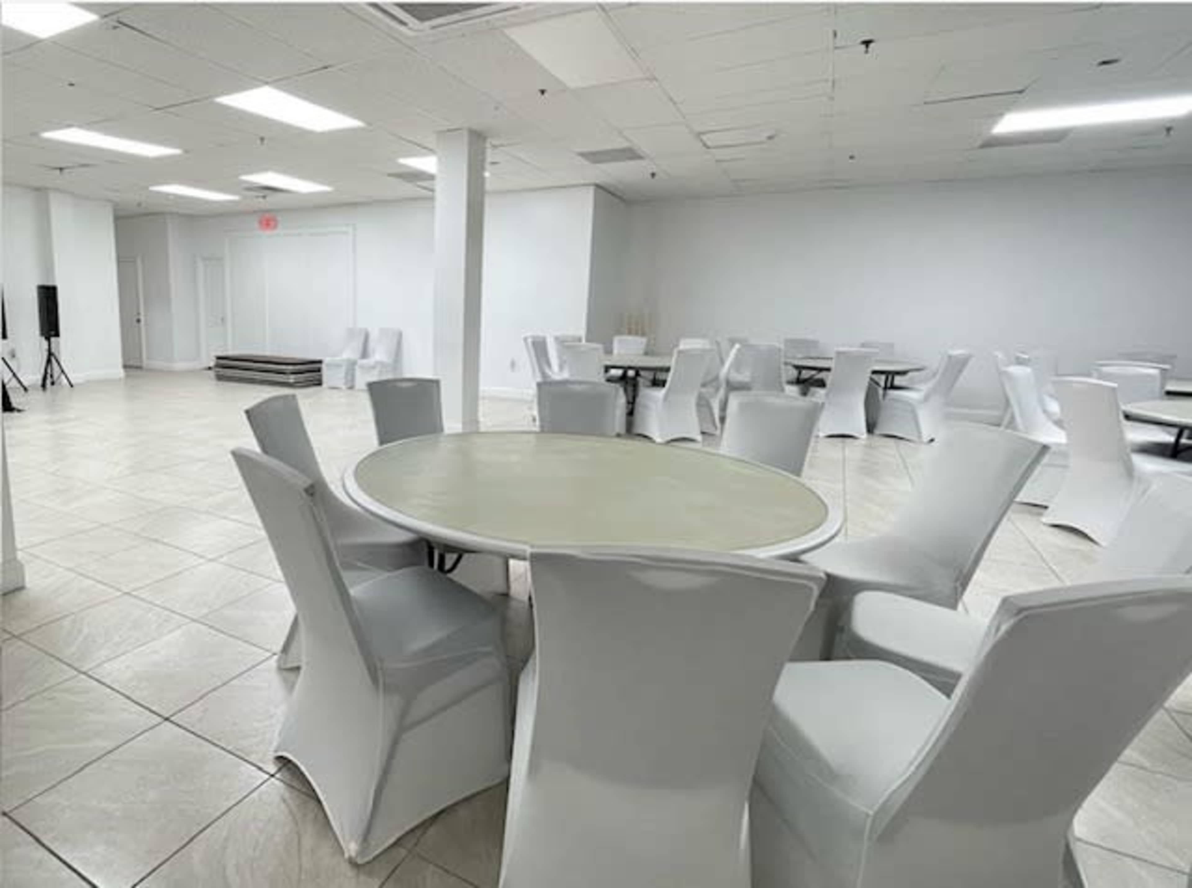 The image shows a spacious, empty room with several round tables surrounded by white chairs.