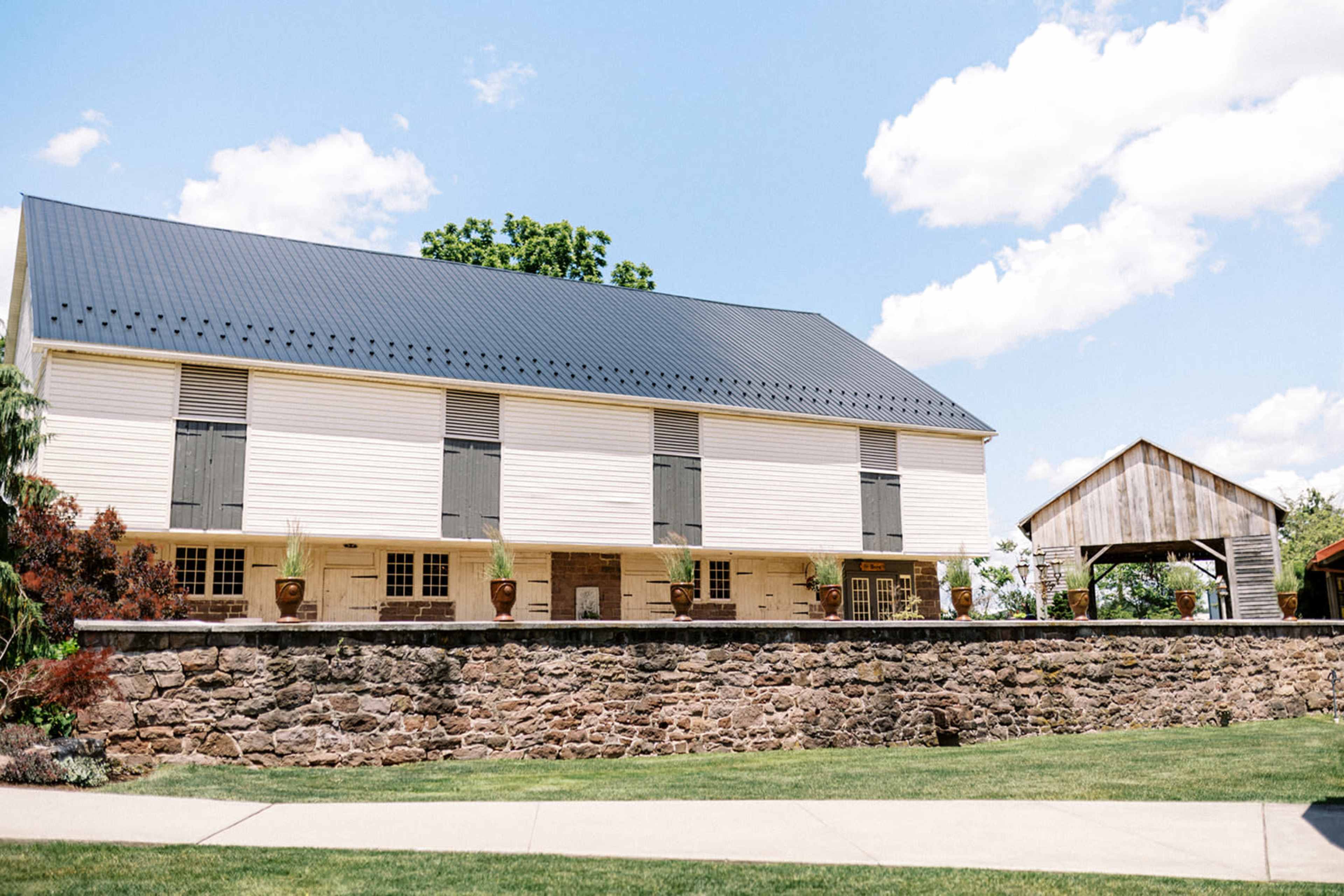 A large, white barn with a black roof and several windows stands next to a smaller, rustic structure, all set against a clear sky.