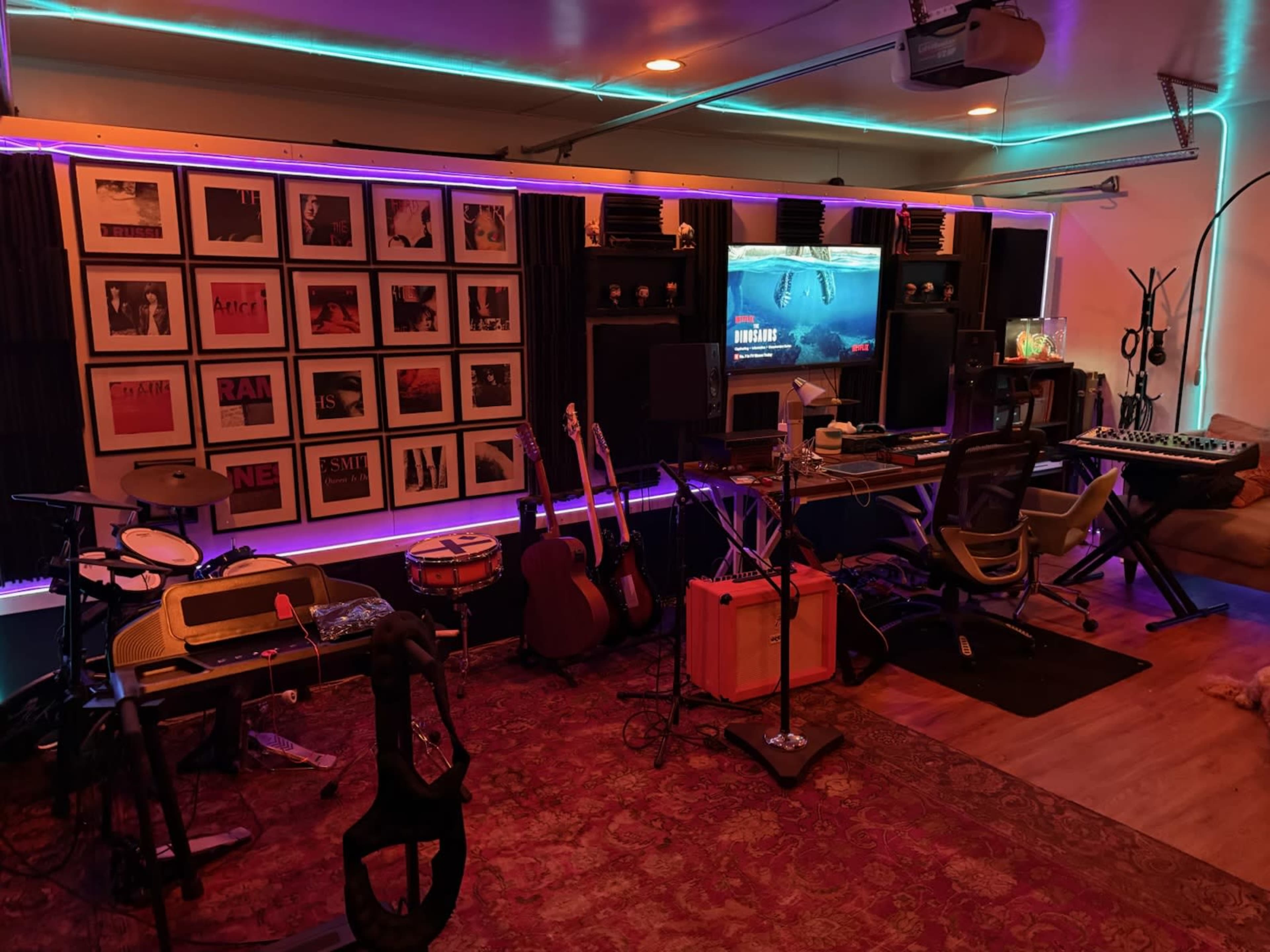 The image shows a home music studio featuring various instruments, acoustic panels, a large screen, and a colorful LED strip illuminating the workspace.