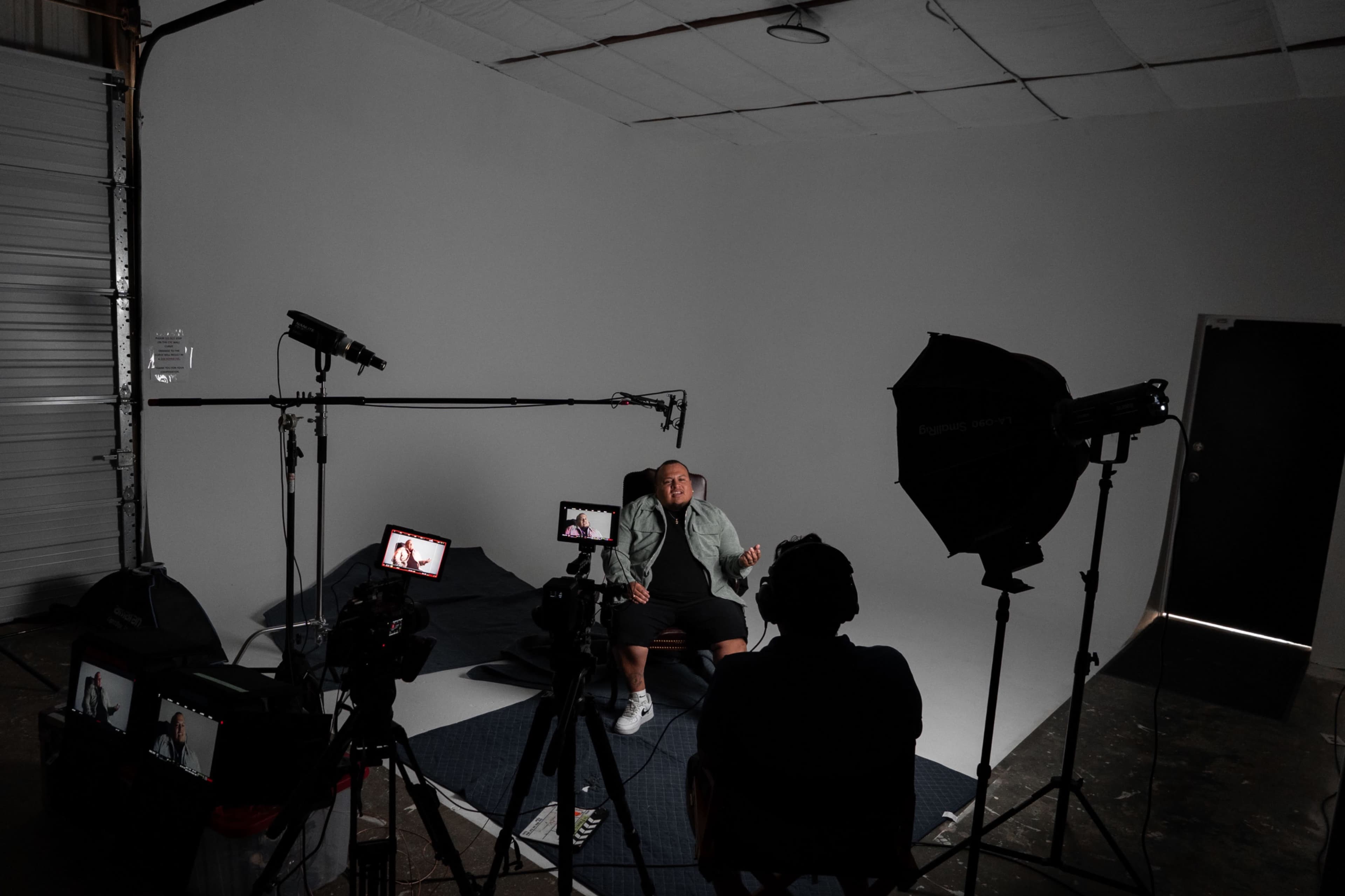 A person sits casually on a stool in a studio setup with multiple cameras and lighting equipment.