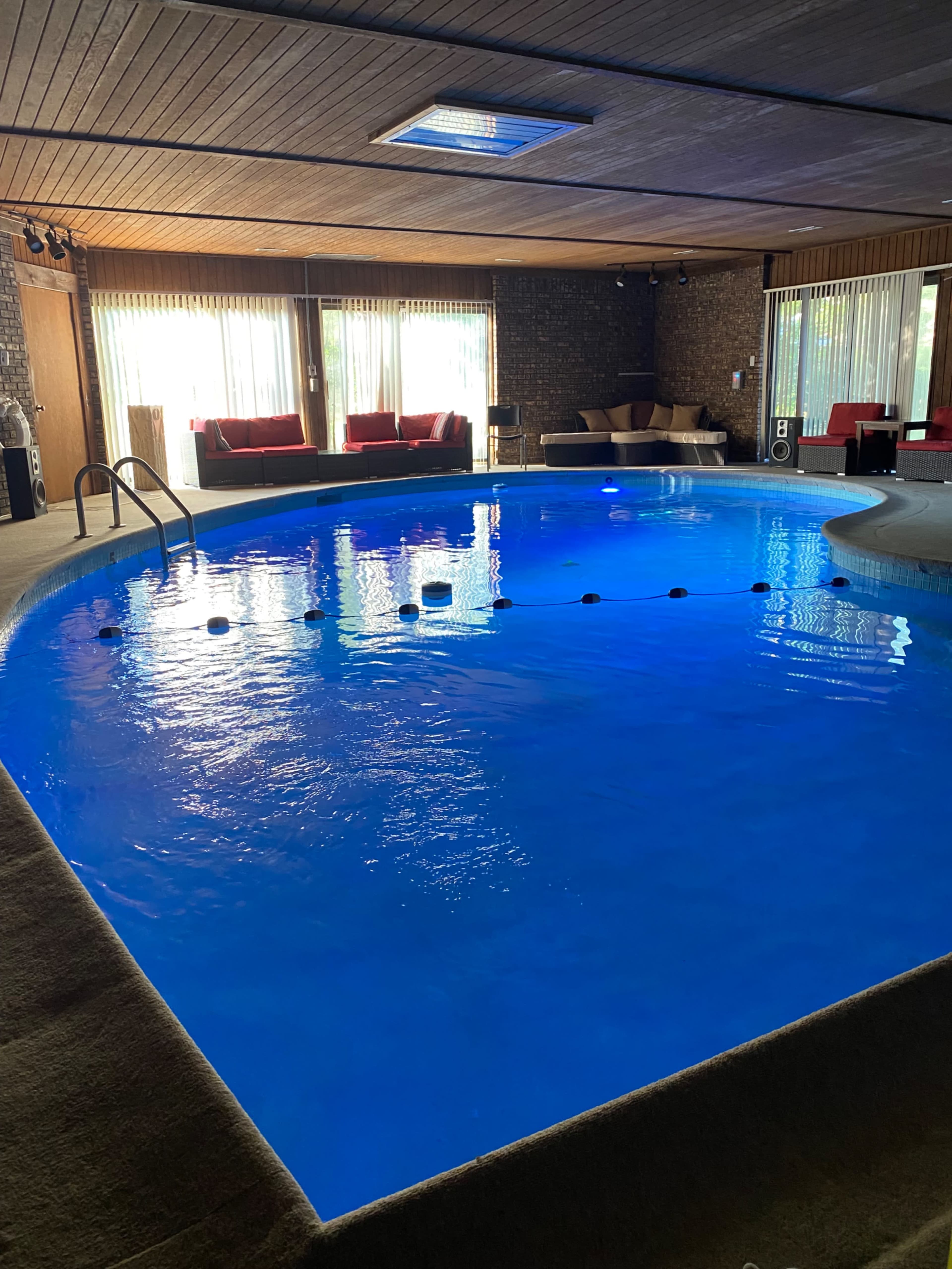 An indoor pool area featuring a blue lit pool, with lounge seating and large windows allowing natural light.