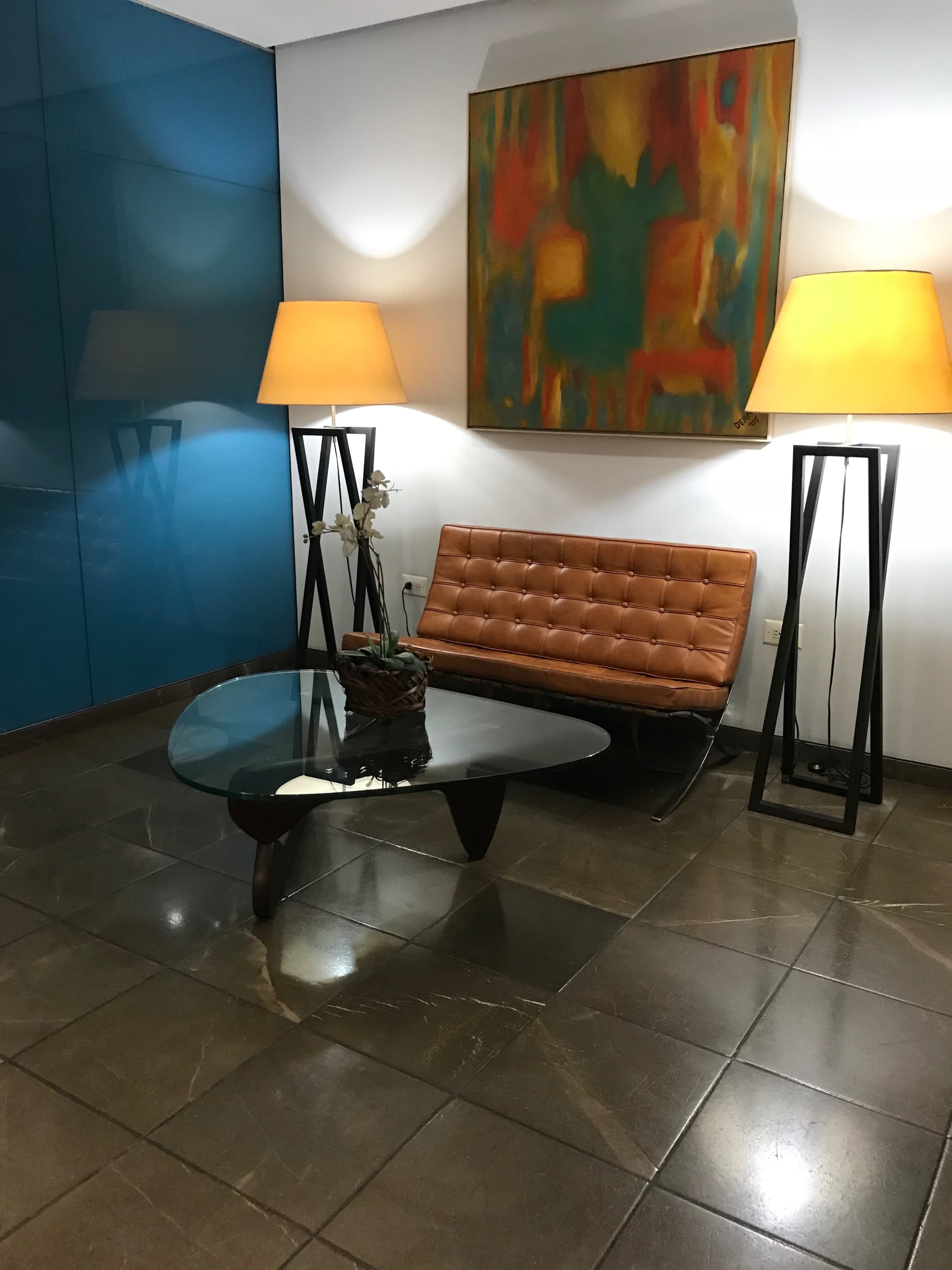 A modern waiting area features a leather sofa, a glass coffee table, two floor lamps, and an abstract painting on the wall.