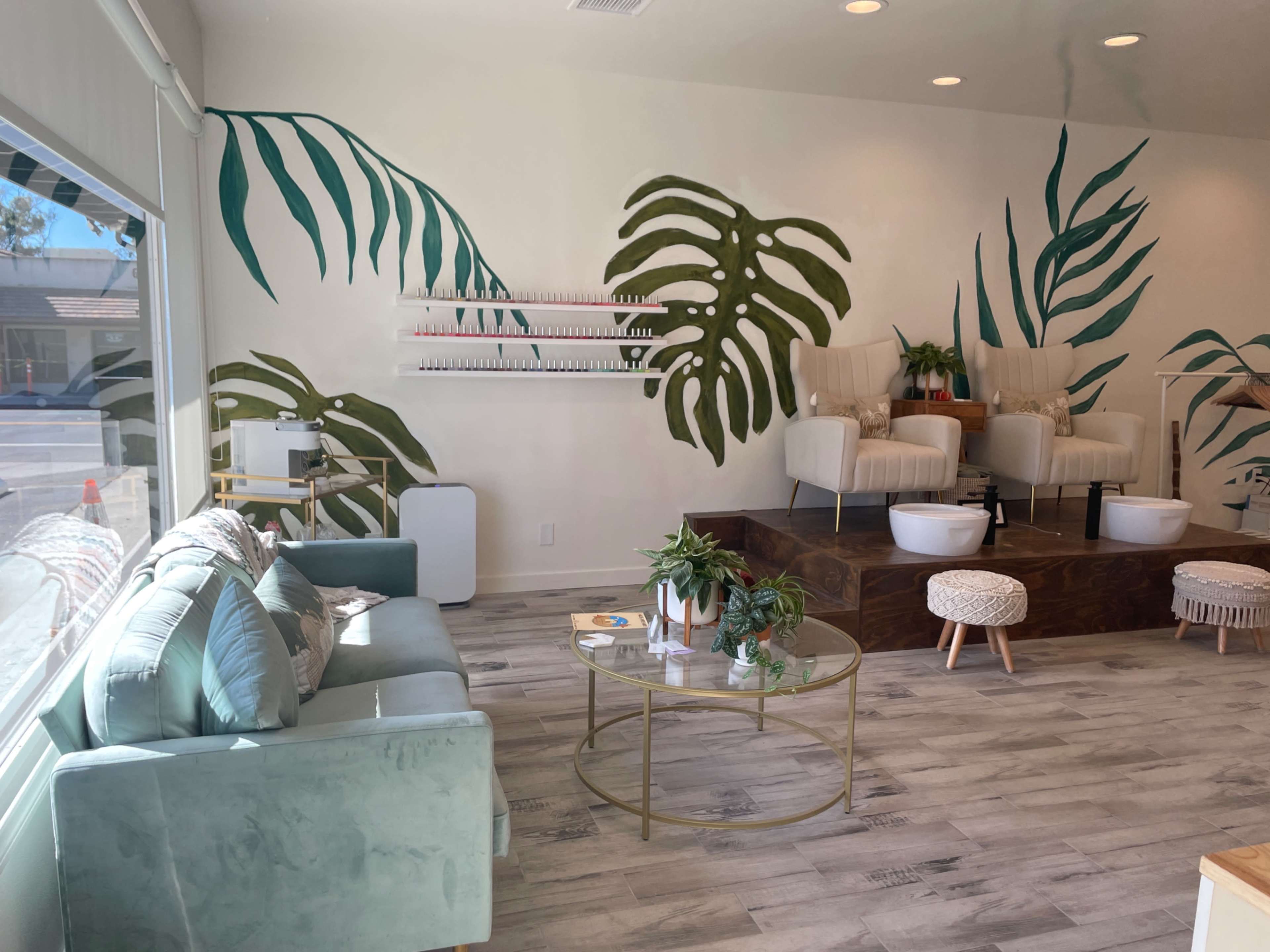 The interior of a nail salon features green leaf murals on the walls, a seating area with a light blue couch, and two white pedicure chairs positioned on a raised wooden platform.