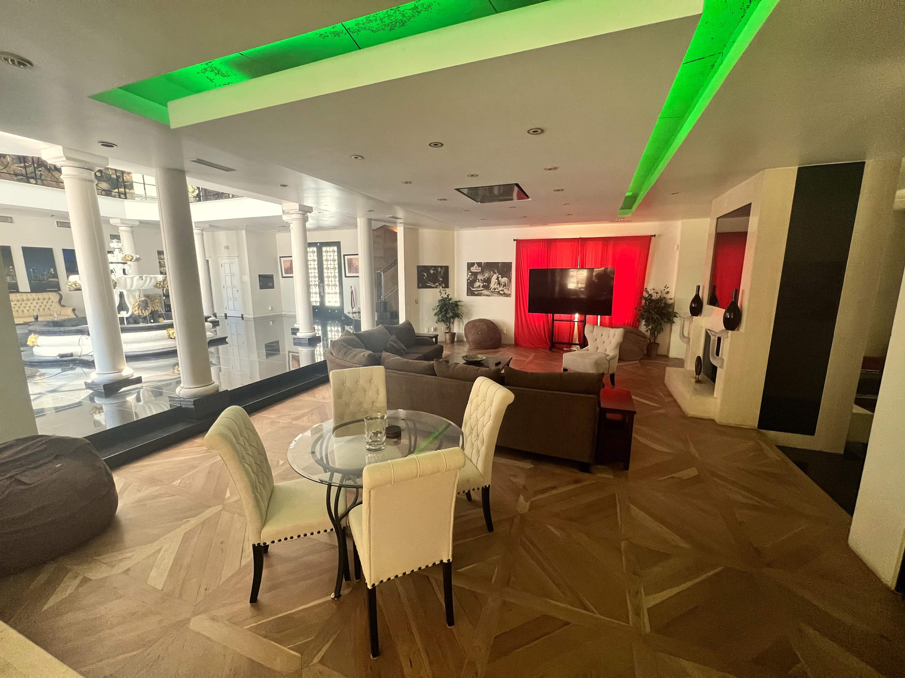 The image shows a spacious living area with modern furniture, including a round glass dining table and a large screen against a red backdrop.