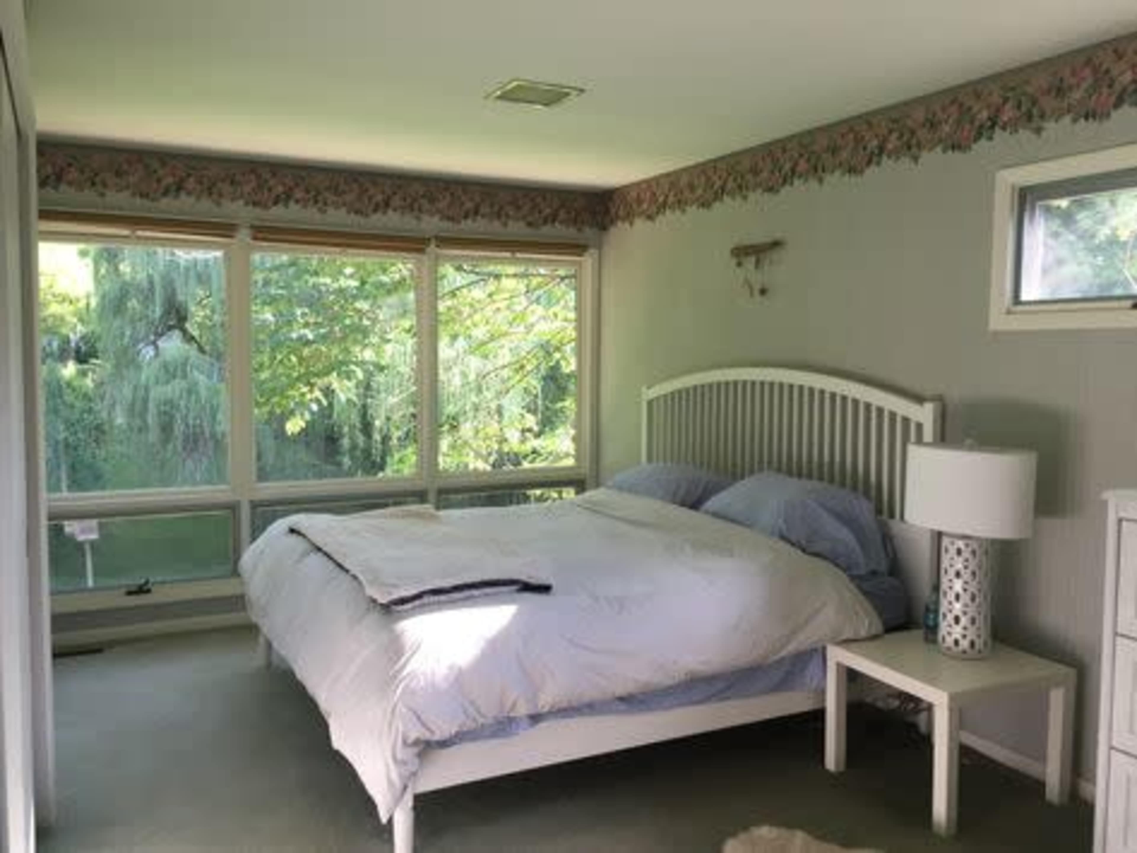 A bedroom features a bed with light blue bedding, a small bedside lamp, and large windows overlooking greenery.