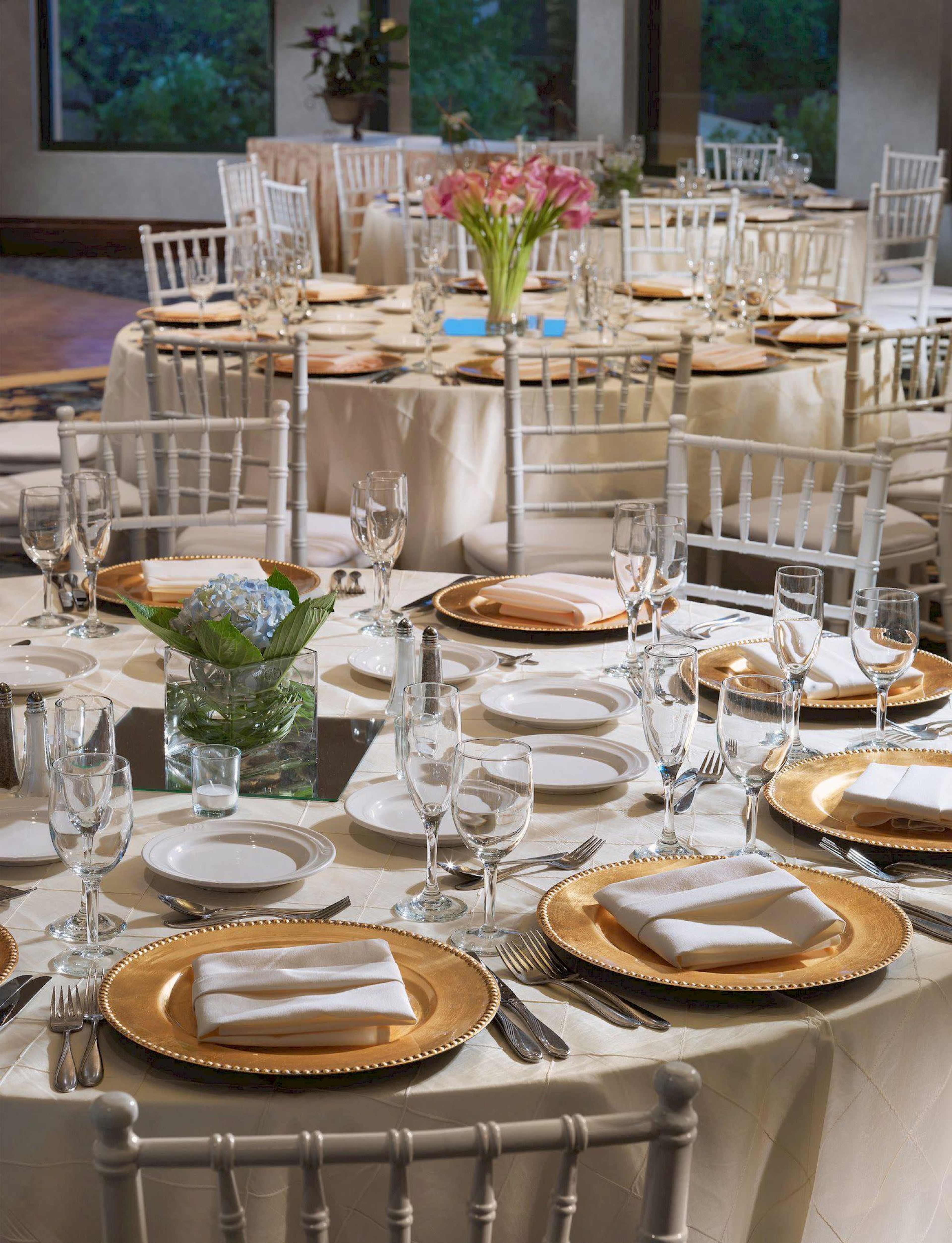 The image shows elegantly arranged dining tables with white linens, gold chargers, and glassware in a well-lit event space.