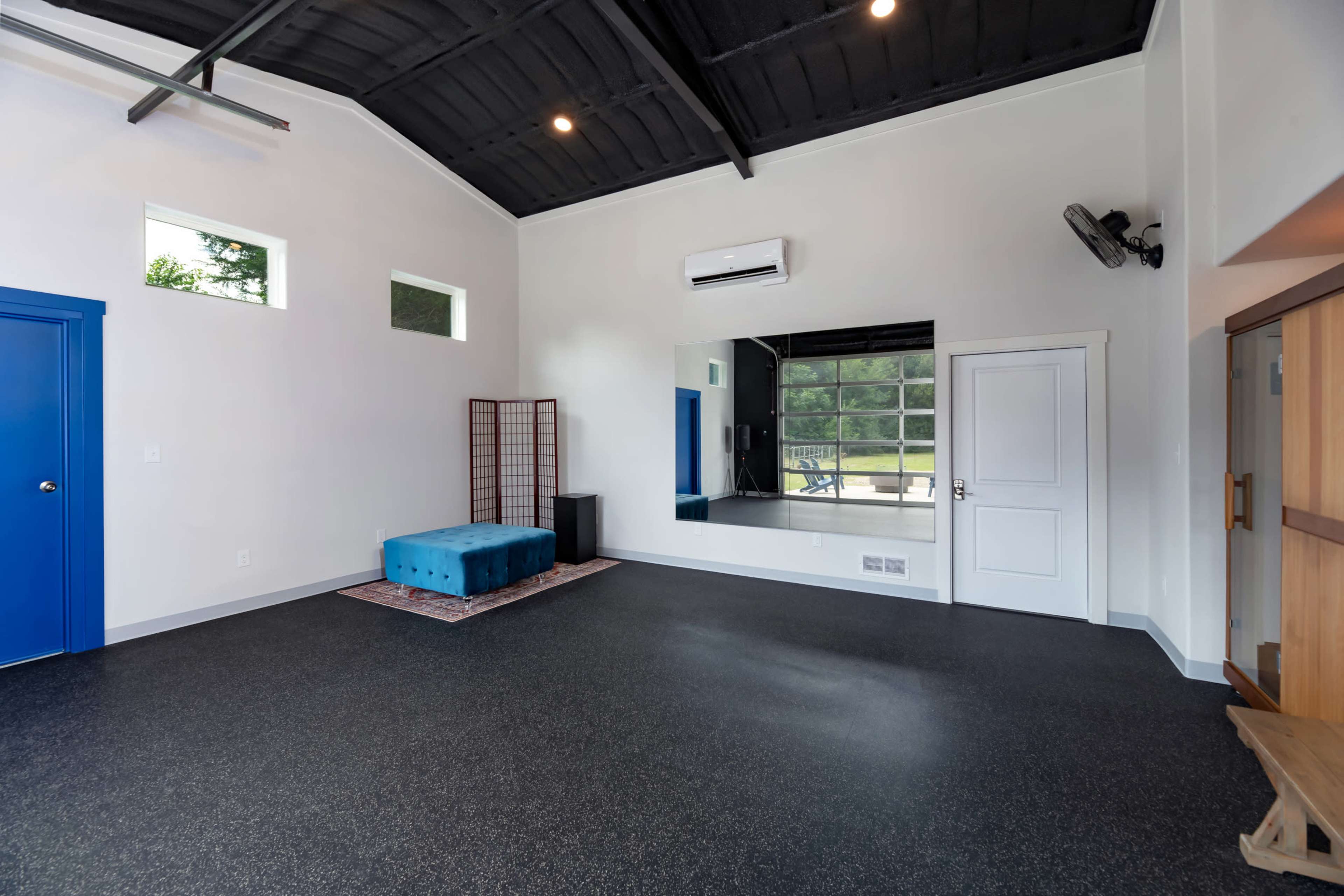 The image shows an empty room with a high ceiling, featuring a blue bench, a large mirror, and a door, all against a backdrop of black flooring and white walls.
