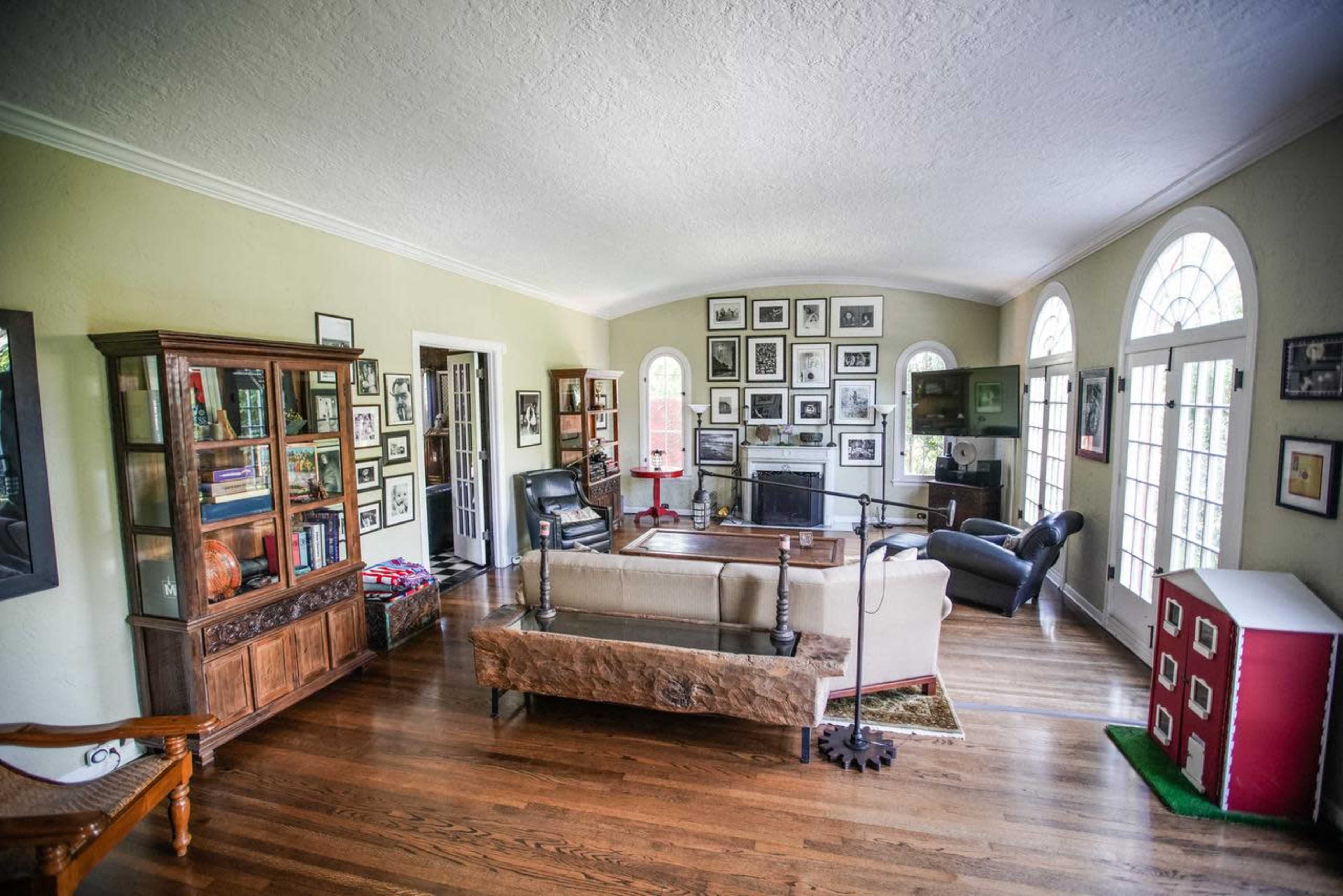 A spacious living room features large windows, wooden floors, a variety of framed photographs on the walls, and a combination of modern and traditional furniture.