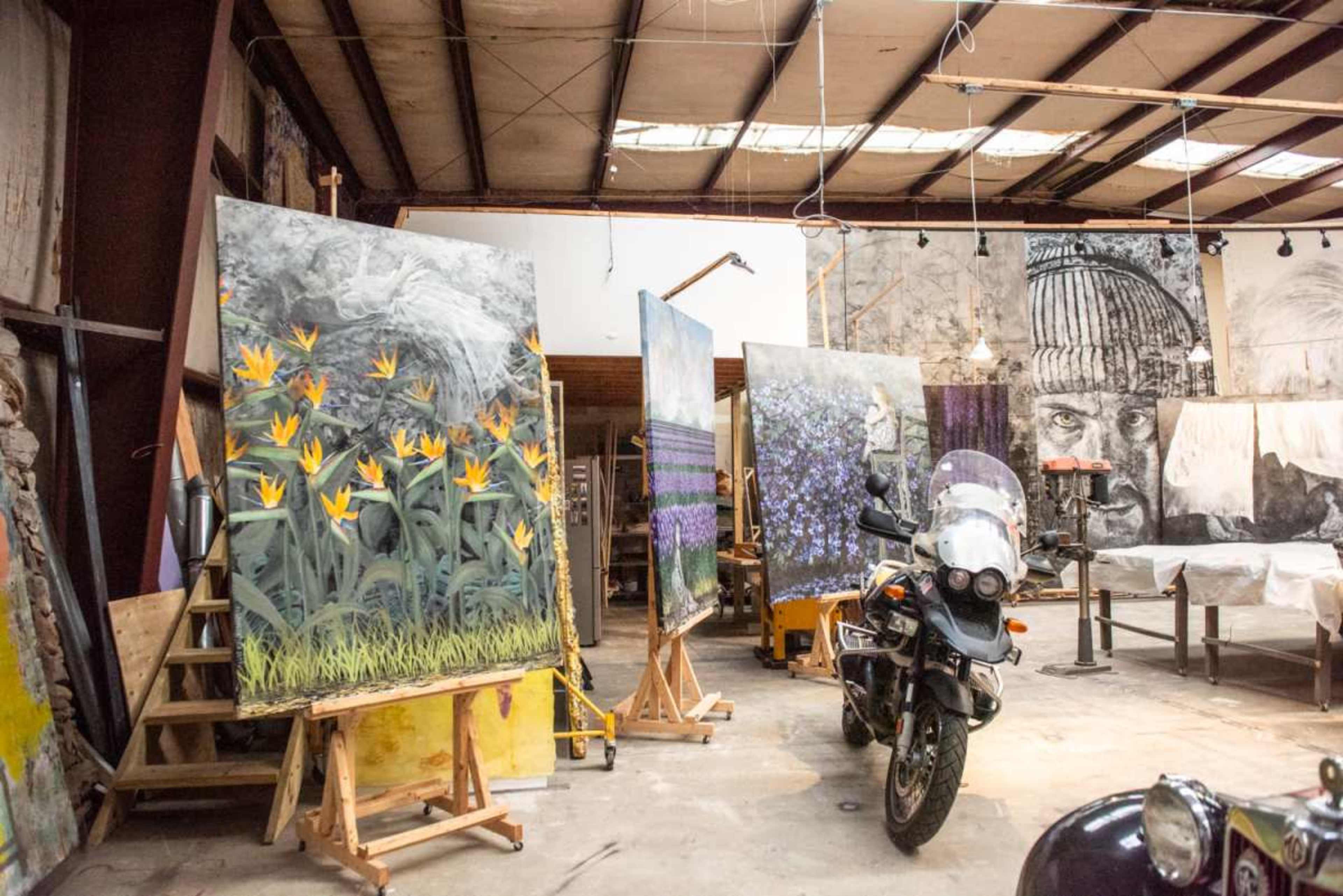 An art studio with several large paintings on easels, a motorcycle parked in the foreground, and various artworks displayed around the room.