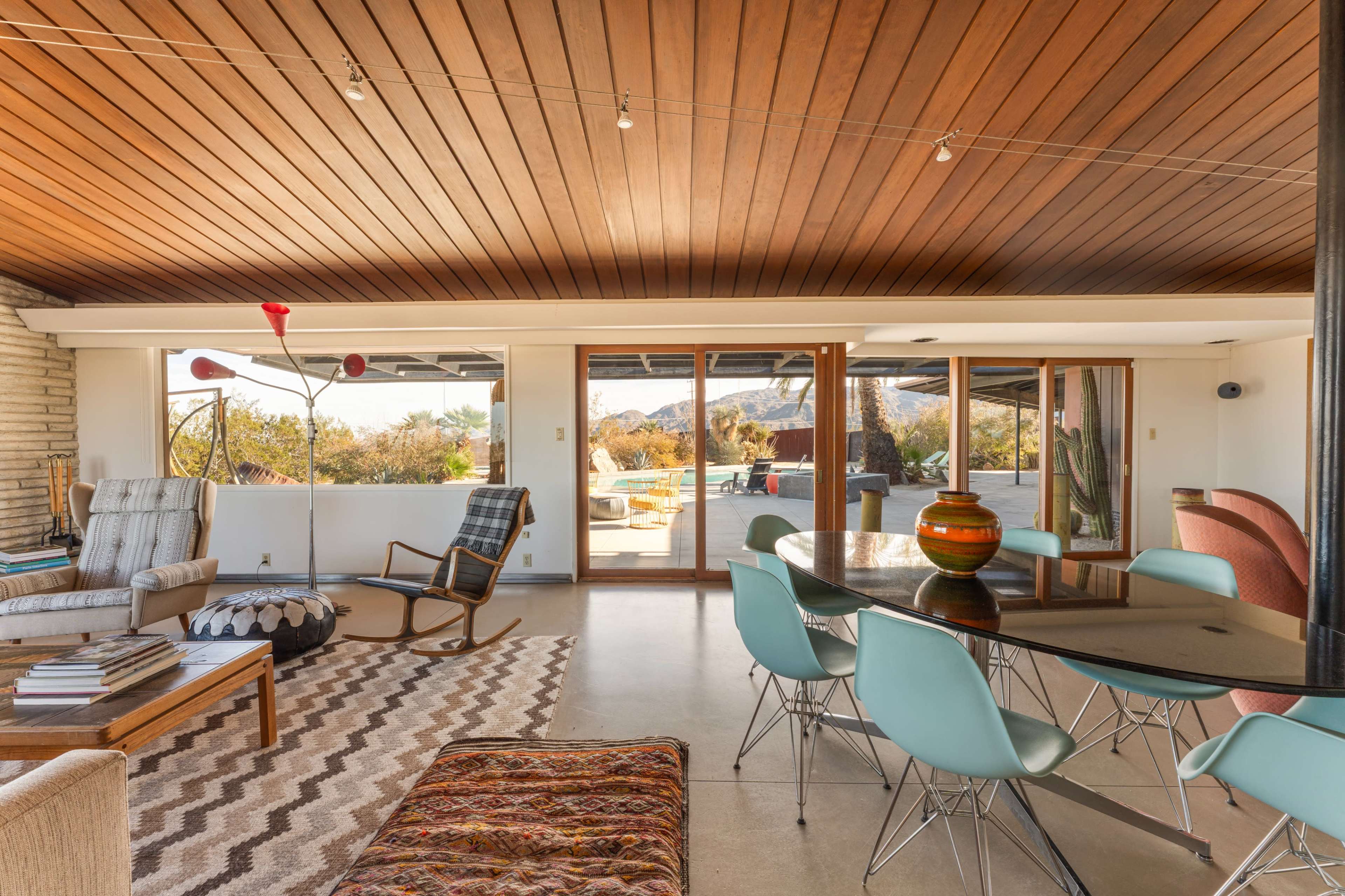 The image shows a spacious indoor living area with a wooden ceiling, large windows overlooking an outdoor patio, and modern furniture including a dining table with turquoise chairs and a patterned rug.